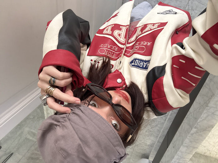A high-angle, close-up image depicts an individual, likely female, engaged in a self-portrait. The subject wears a white, red, and black racing-style jacket featuring embroidered patches, including "CHARIOT" on the left chest and "PION READY" on the right. Dark brown hair is visible beneath a light grey fabric cap, the brim of which is held by the subject's left hand. Dark, oval-shaped sunglasses are positioned on the face, with the right hand holding them slightly lowered, revealing the subject's eyes looking directly at the camera. Red lipstick is applied. Multiple rings, including stacked bands and a prominent silver ring, adorn the fingers of the right hand. The background consists of a light-colored marble tiled surface with grey veining, appearing as a floor or lower wall, which transitions to a plain white upper wall or ceiling. The specific location within Barcelona, Spain, is not discernible from the visual content.