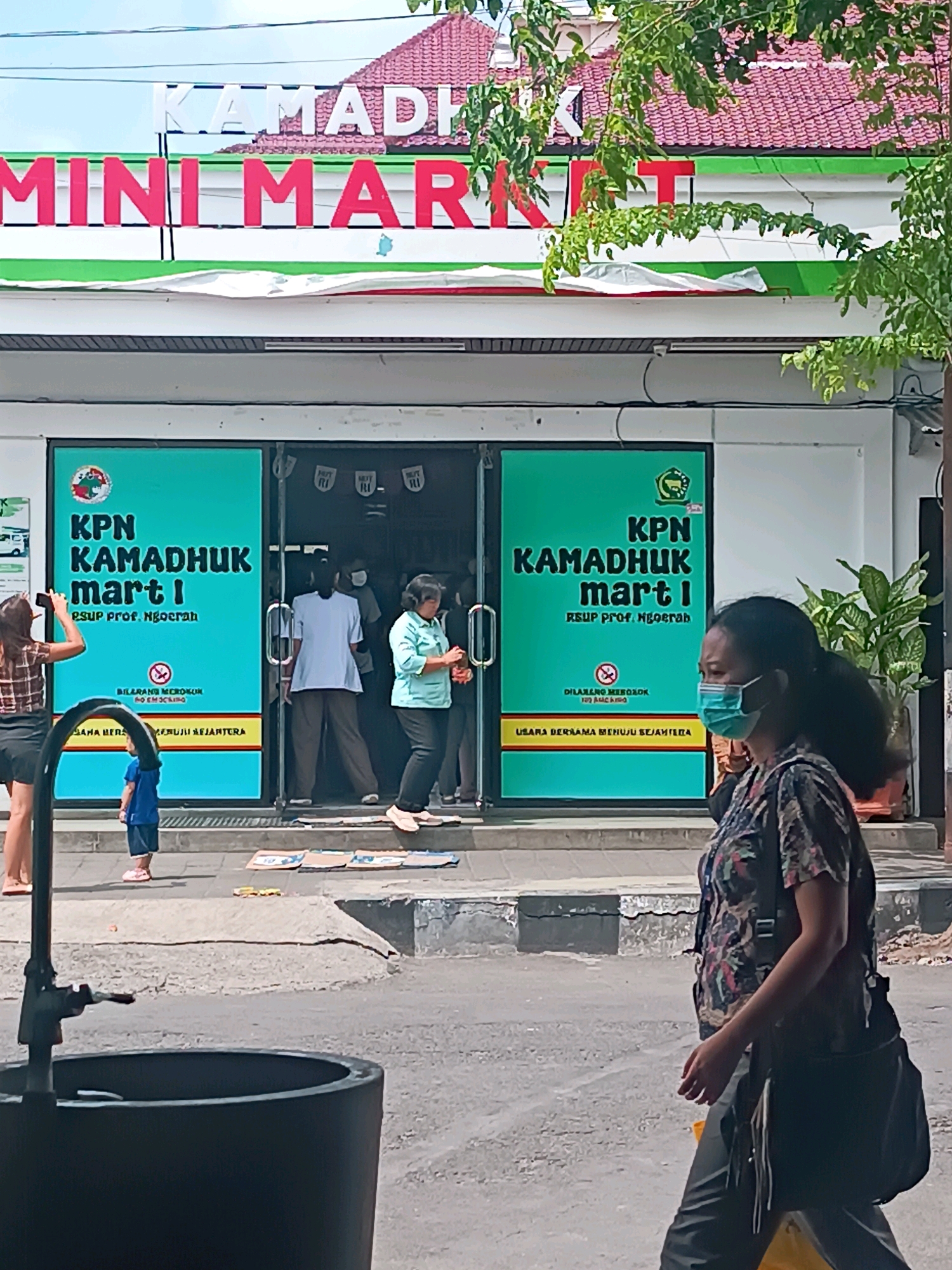 The image captures the exterior of a "Kamaduk mini market" in Denpasar, Indonesia. A large green sign on the storefront reads "KPN KAMADHUk mart 1 RSUP PROF. NGOERAH" and features a smaller "DILARANG MEROKOK" (No Smoking) symbol. Two women are visible; one on the left is taking a photo with her phone, and another on the right, wearing a face mask, walks past the storefront. A young child in a blue shirt and pink shoes stands near the entrance. Inside the mini market, several people are queuing. The building has a red-tiled roof and green and white trim. The street in front of the market is paved, with a dark-colored outdoor bin or planter in the foreground. Cardboard mats are laid out on the sidewalk in front of the entrance.