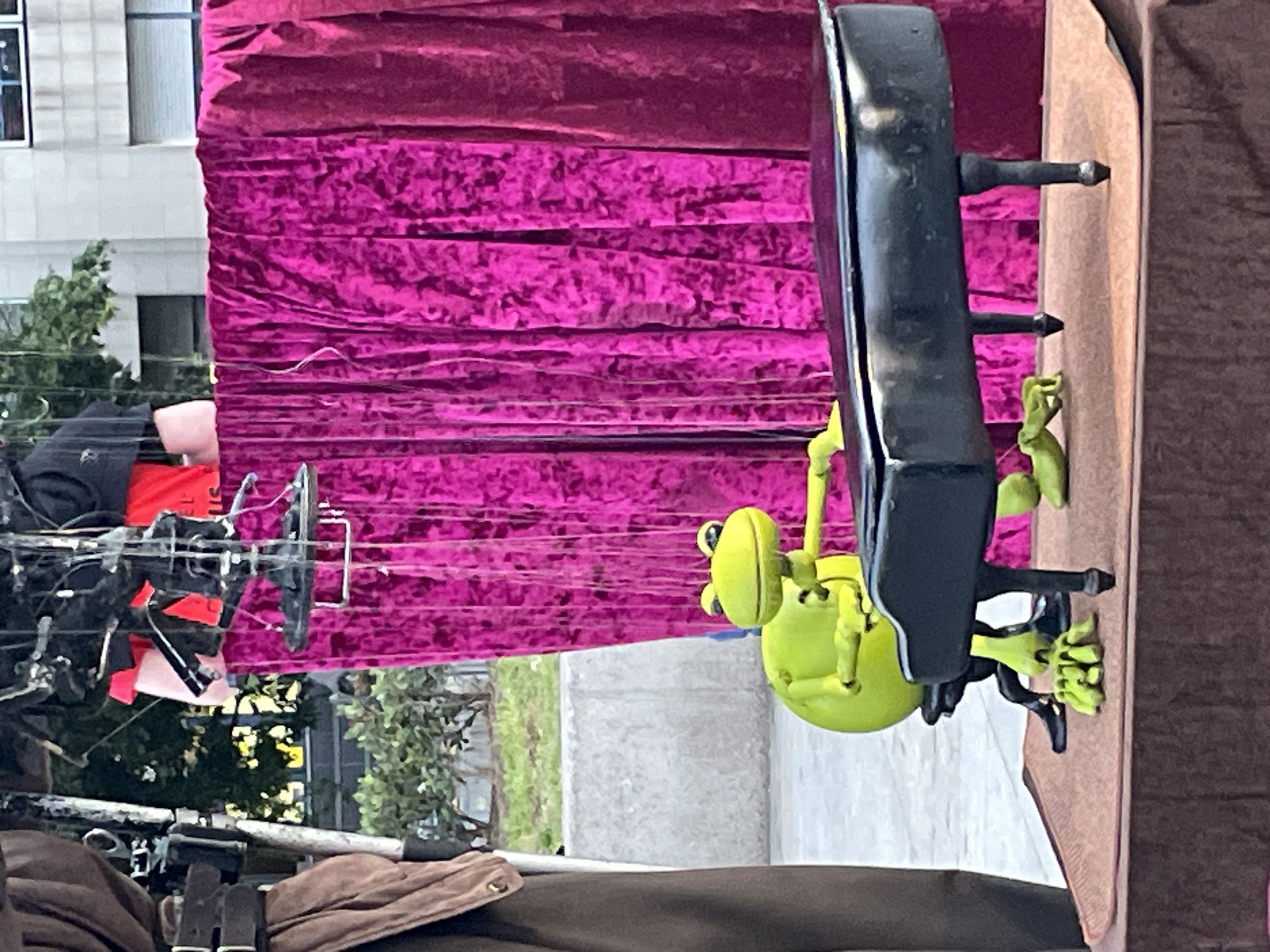 The image depicts a street performance setup. A hot pink velvet curtain serves as a backdrop. To the right, a puppet of a green frog plays a miniature black piano. Wires and rigging are visible, indicating the puppet is controlled by a performer. The scene appears to be outdoors in an urban setting, with buildings and greenery visible in the background. Based on the urban architecture and plant types visible, the location is most likely in a major city in Europe. However, there is insufficient information to confirm the location is in Barcelona, Spain.