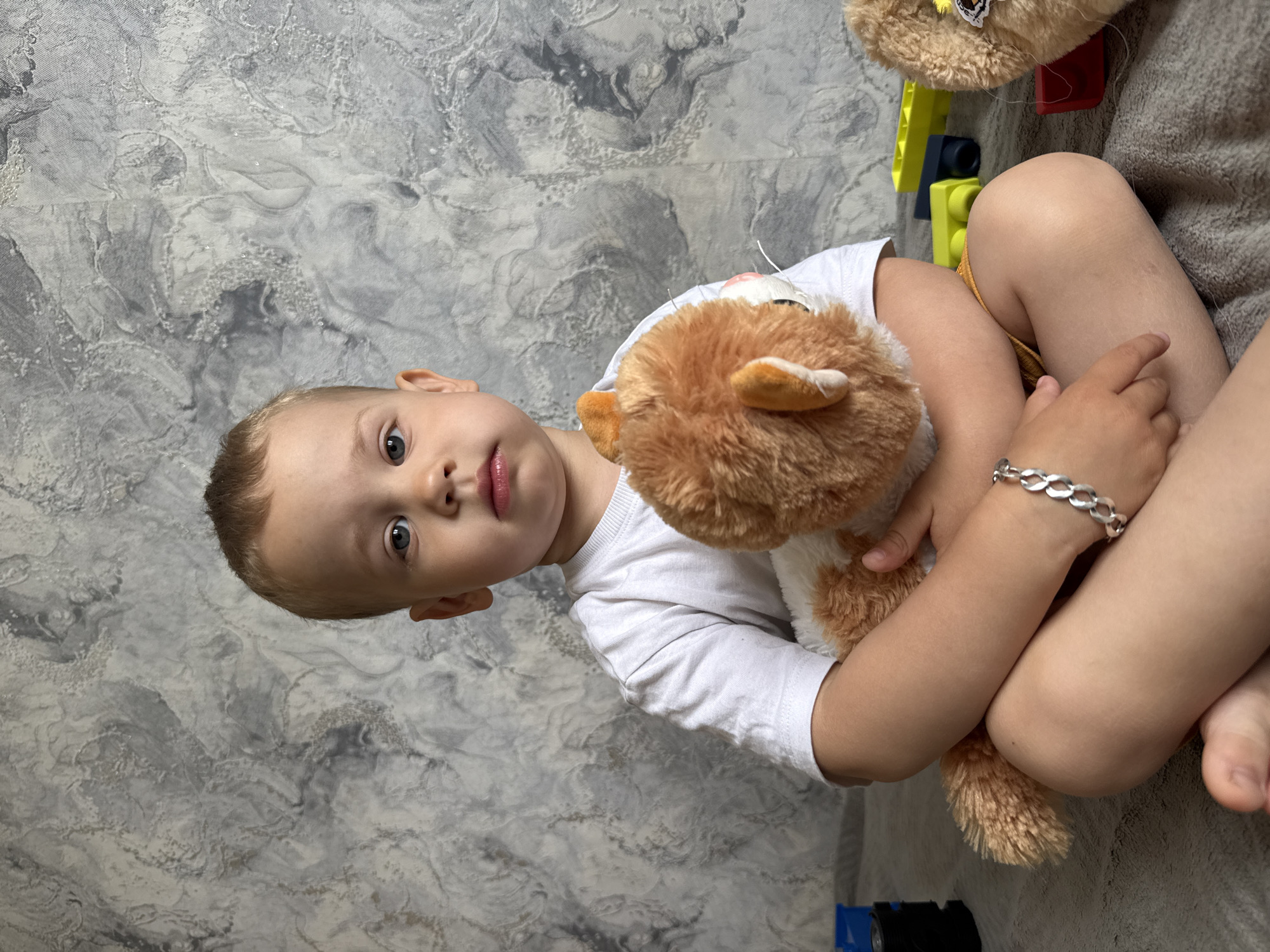 A young, fair-skinned child with short blonde hair is holding a stuffed orange and white cat. The child is wearing a white t-shirt and a silver bracelet. The child is sitting with legs crossed, with a gray blanket visible underneath. Behind the child is a gray and white textured wall. There are also yellow and blue toy blocks visible in the background. The location is Rusanovo, Russia.