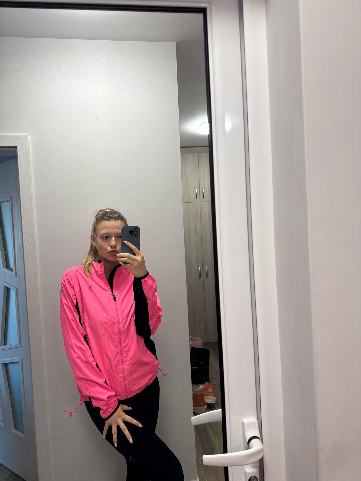 A woman is taking a mirror selfie in a room. She is wearing a bright pink jacket with black accents and black pants. She is holding a smartphone in her right hand and has her left hand on her hip. Her hair is pulled back in a ponytail. In the reflection of the mirror, a closet with wooden doors and a light fixture can be seen. The room appears to be a hallway or a dressing area, with a white door on the right side of the image. The location is not specified as Sofia, Bulgaria.