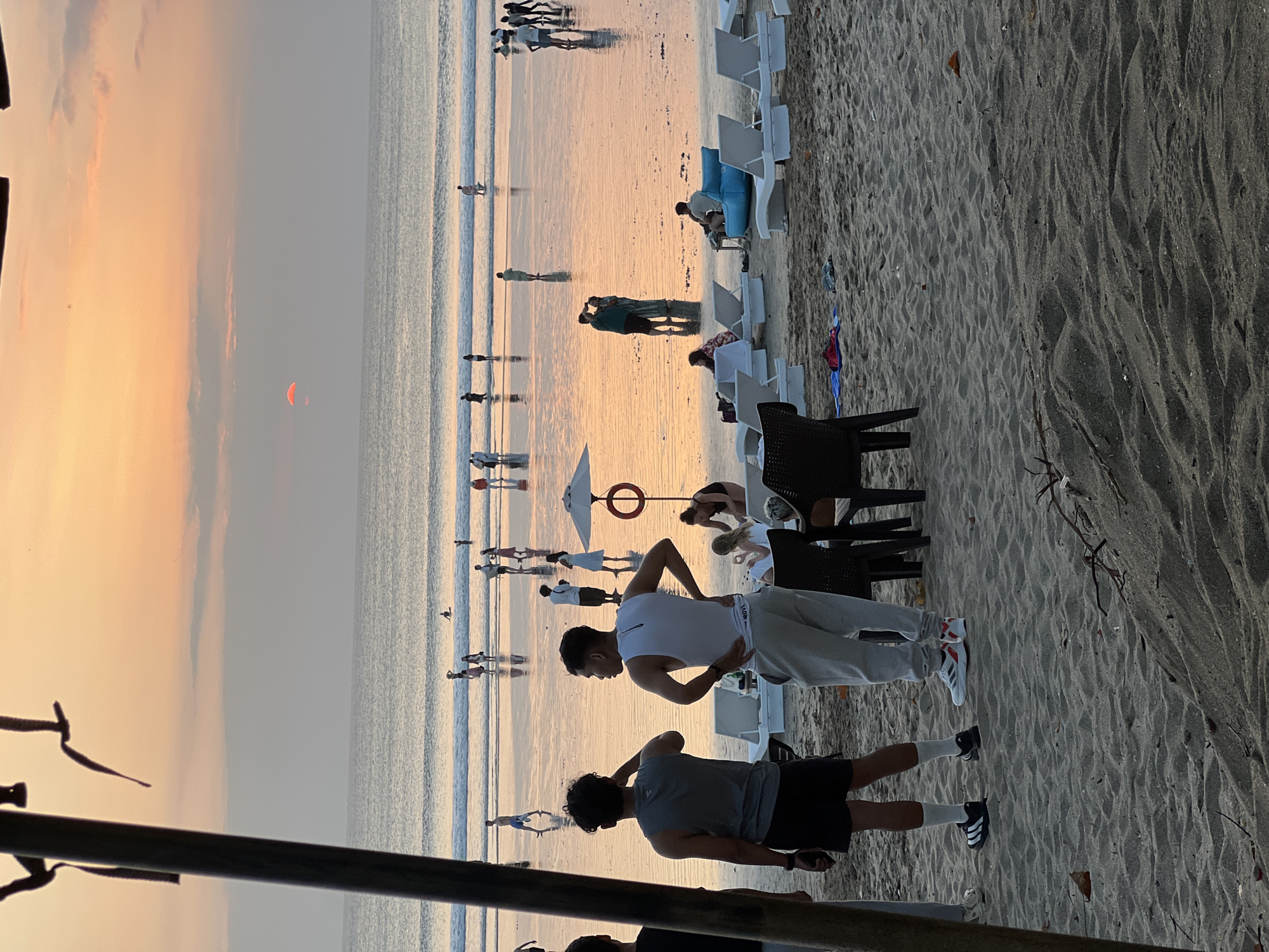 A group of people are gathered on a sandy beach at sunset. The sky is a warm orange and pink, with the sun partially visible on the horizon. Waves are gently rolling onto the shore, and the wet sand reflects the colors of the sky. Several beach chairs are set up on the sand, some occupied by people lounging. In the foreground, two men are standing and looking out at the ocean. In the background, more people are walking along the water's edge and in the shallow surf. The scene appears to be in Kuta, Bali, Indonesia, a popular tourist destination known for its beaches and sunsets.