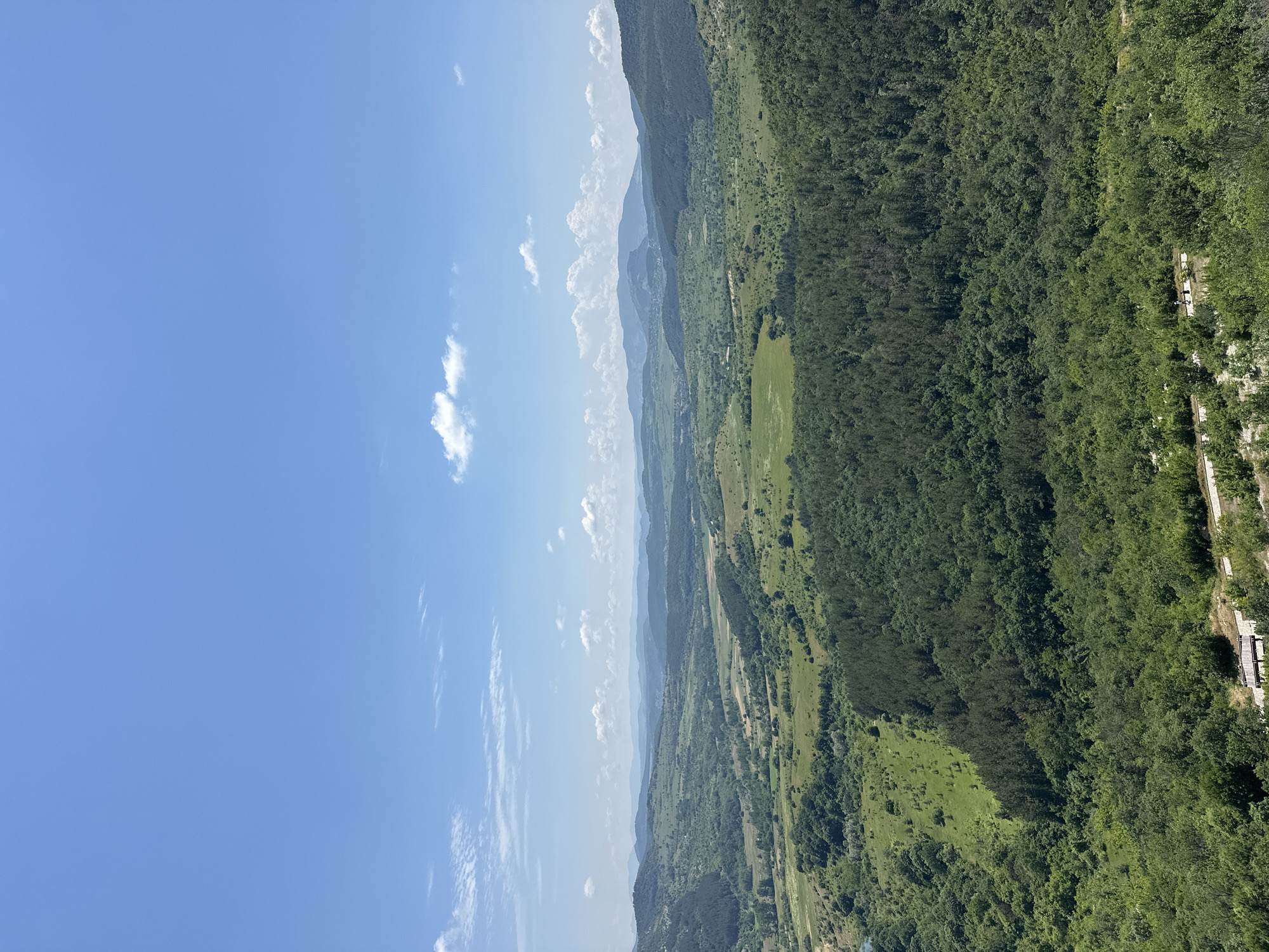 This is a wide-angle landscape photograph depicting a vast, verdant valley and rolling hills in the vicinity of Gorna Krepost, Bulgaria, under a clear, bright blue sky with scattered white clouds.

The upper third of the image is dominated by the light blue sky, which features several wispy cirrus clouds and a few smaller, puffier cumulus clouds, primarily in the lower left quadrant of the sky.

The horizon line reveals multiple layers of distant, hazy blue-gray mountains and hills, indicating significant depth in the landscape. The midground is a broad expanse of lush green terrain, characterized by an irregular mosaic of dense, darker green forested areas and lighter green open fields or meadows. Some fields appear to be cultivated or managed, while others are wilder. A slender body of water, possibly a river or stream, is visible in the valley towards the far left. A small cluster of buildings can be faintly discerned in a distant valley, nestled among the hills.

In the immediate foreground, dense, dark green deciduous and coniferous forest dominates, suggesting the photo was taken from a high, forested vantage point. An unpaved, light-colored path or road winds across the lower right portion of the image, partially obscured by foliage. On this path, two small human figures are discernible, appearing to walk from left to right. Further along the path to the right, a wooden bench or similar seating structure is partially visible beneath the trees. The figures appear to be in transit, with no discernible direct interaction between them. The overall environment is natural and rural, under clear, sunny daytime conditions.