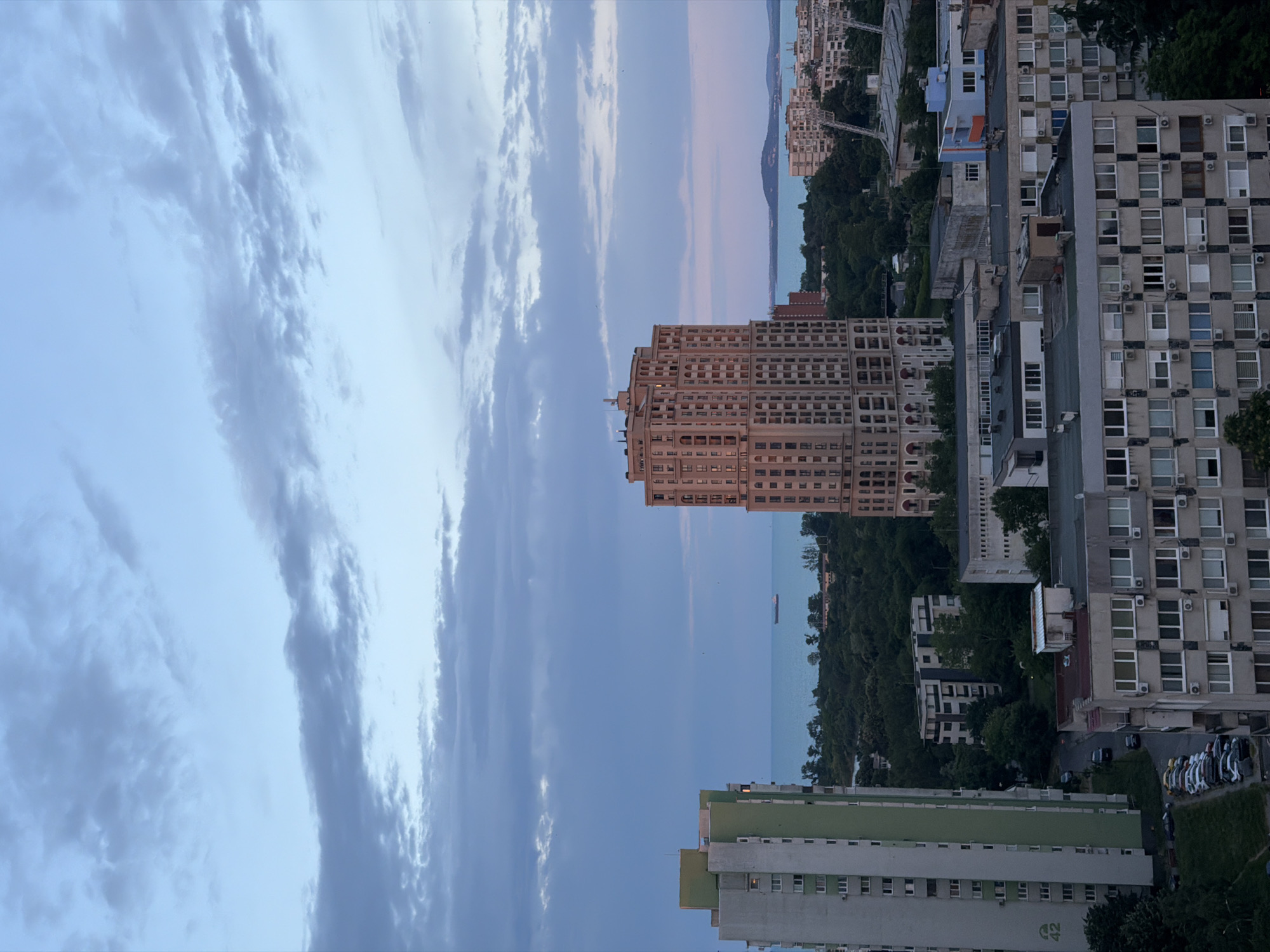 The image captures a panoramic view of the city of Burgas, Bulgaria, at dusk. In the foreground, a tall, modern apartment building with a distinct ornate facade dominates the right side of the frame. To its left, a taller, more utilitarian apartment building with a green vertical stripe is visible. The middle ground is filled with a dense canopy of trees, interspersed with various other buildings of different architectural styles and heights, suggesting a mixed urban landscape. In the distance, the Black Sea stretches across the horizon, its calm surface reflecting the soft hues of the twilight sky. A single cargo ship is visible on the water, adding a touch of maritime activity to the scene. The sky above is a canvas of soft blues and grays, with scattered clouds indicating the transition from day to night. The lighting suggests it is either early morning or late evening. The scene is devoid of any visible human figures.