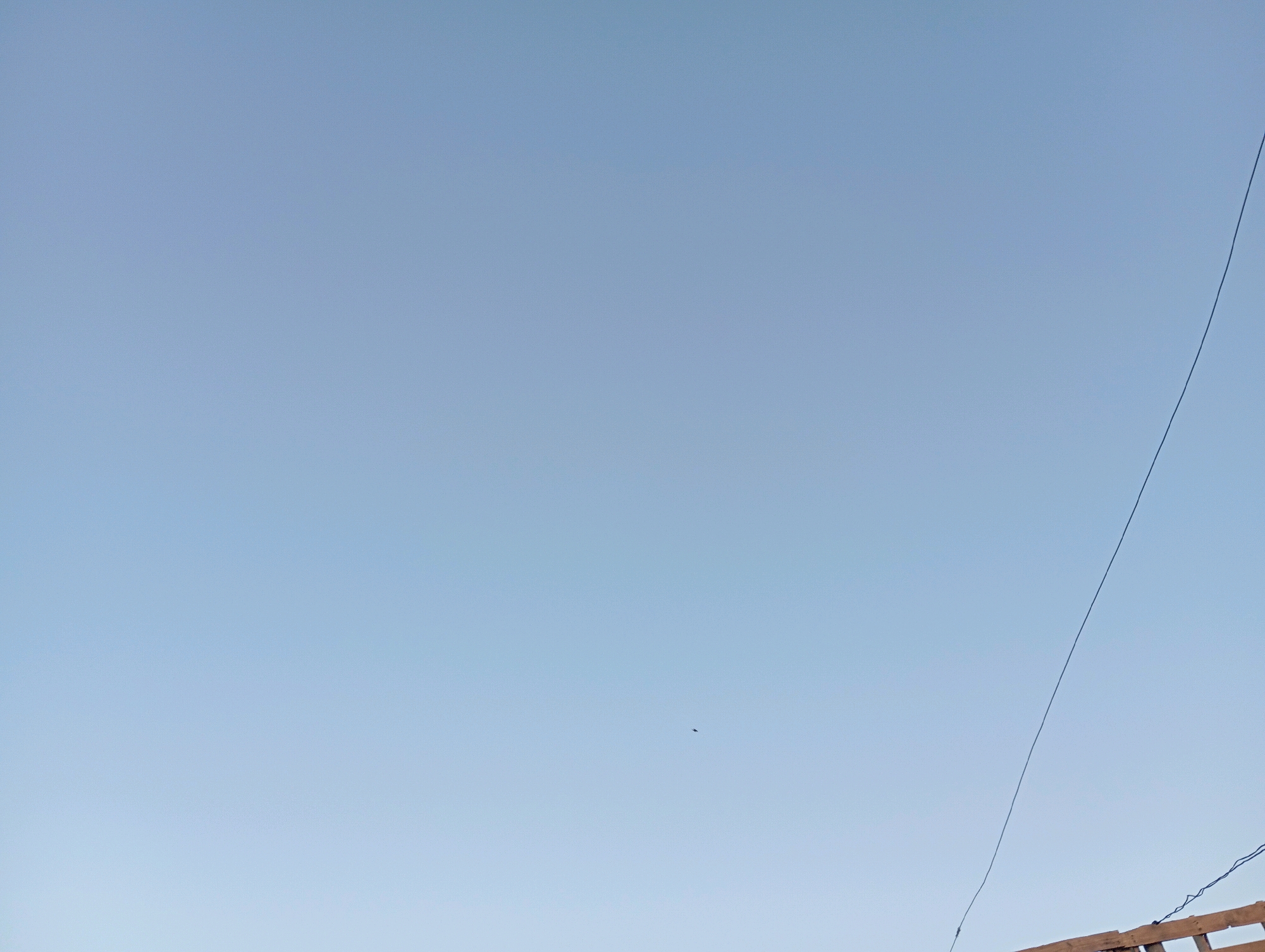 The image shows a clear blue sky above a rooftop structure. A thin black wire stretches diagonally across the upper right portion of the frame, originating from the top edge and disappearing towards the bottom. In the center of the sky, a very small dark speck is visible, which could be a distant bird or an insect. The rooftop appears to be made of wooden planks, with some of the planks forming a lattice-like structure. The overall scene is tranquil and airy, suggesting an outdoor setting in Marrakesh, Morocco.