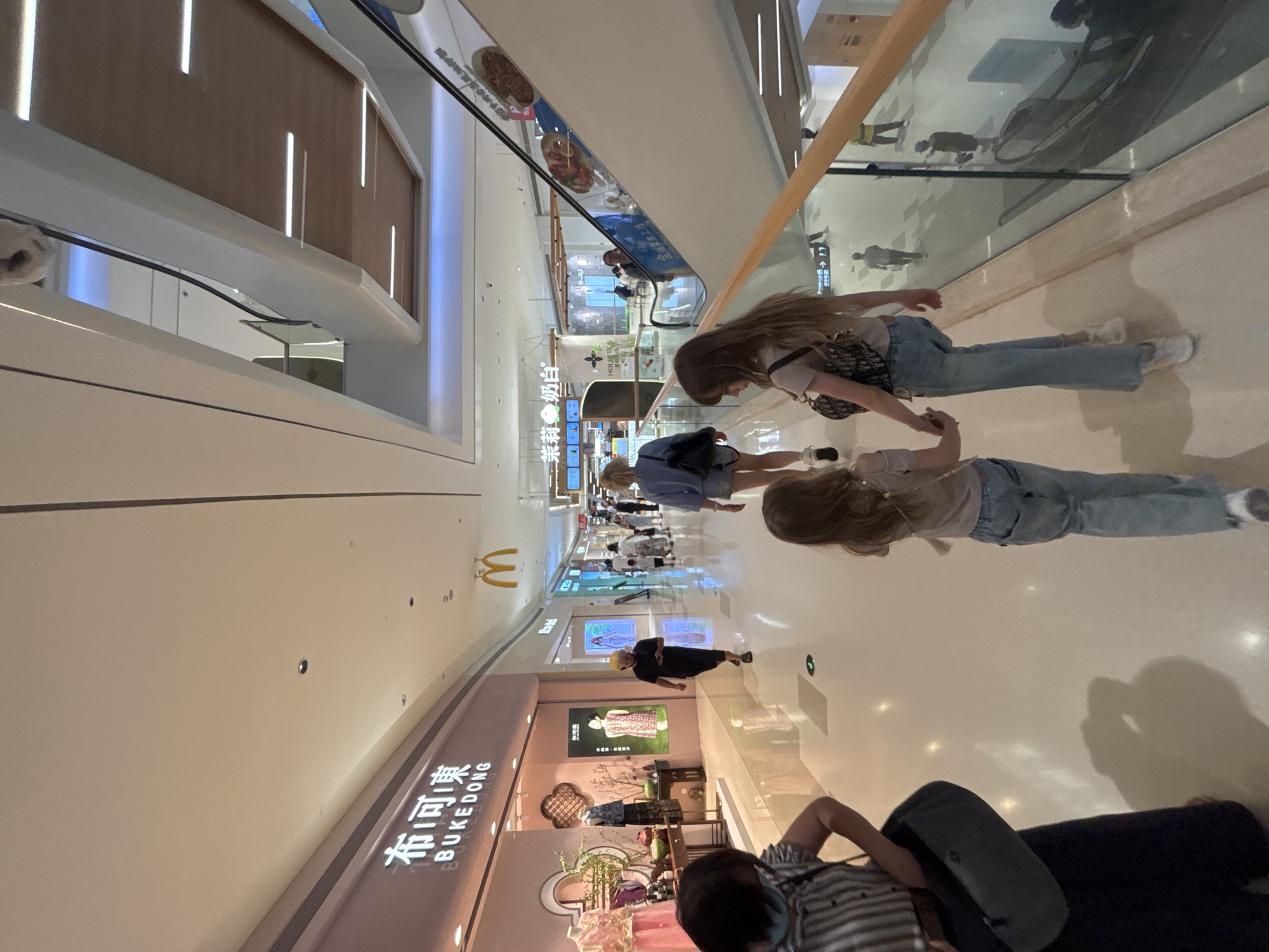 In Tianhe District, China, two young girls are walking hand-in-hand through a brightly lit, modern shopping mall. The girl on the left has long, blonde hair and wears a light-colored t-shirt and denim jeans. The girl on the right has long, brown hair and is wearing a similar outfit, carrying a patterned crossbody bag. They are walking away from the camera on a polished, light-colored floor. In the background, other shoppers are seen walking and interacting within the mall. To the left, a store with the signage "BUKEDONG" is visible, displaying clothing on mannequins. Further in the distance, a McDonald's sign is illuminated. An escalator is on the right side of the frame, with people using it to ascend. The mall's architecture features contemporary design elements with a mix of white ceilings, wooden accents, and ambient lighting.