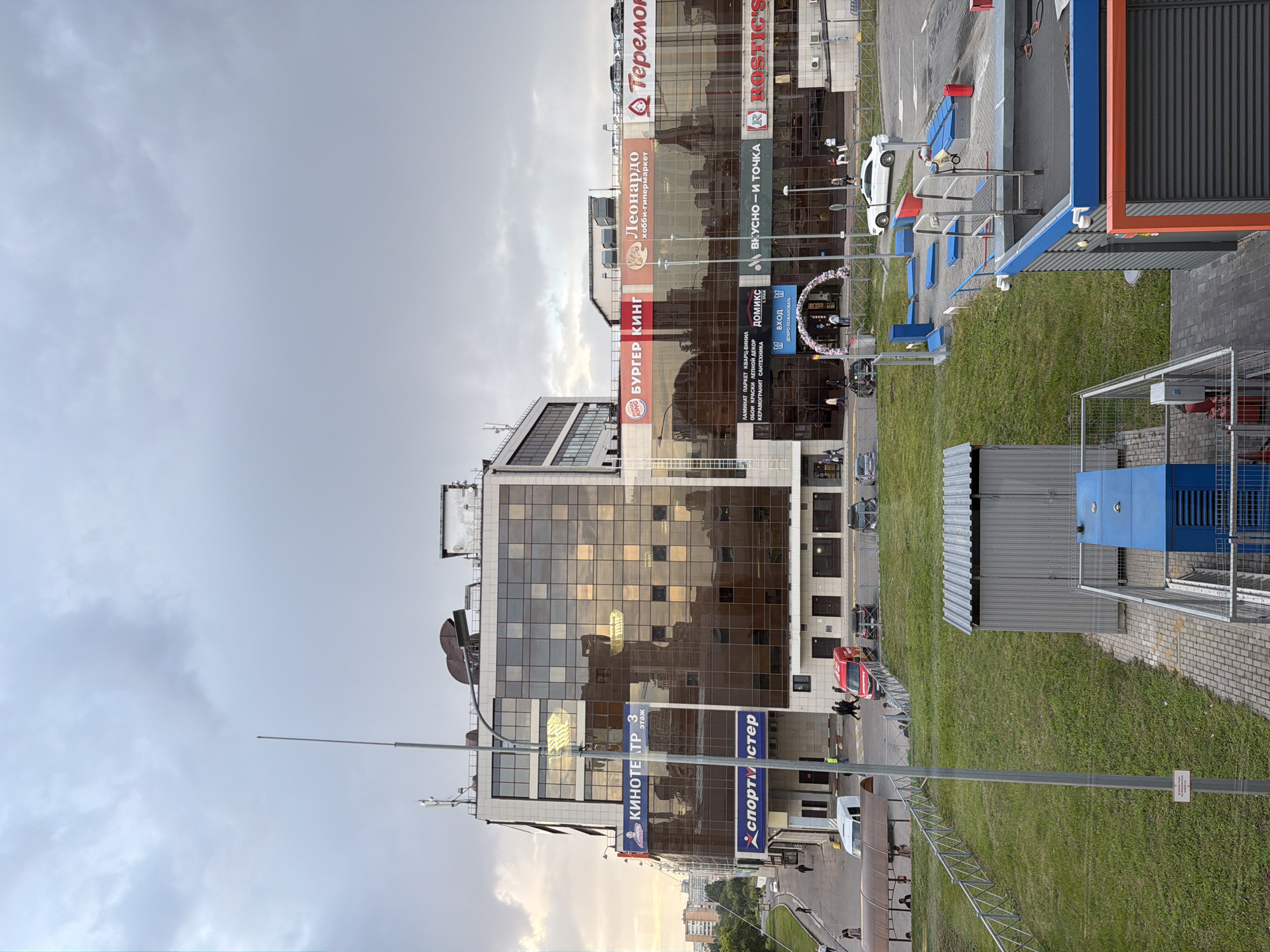 This is an outdoor, eye-level, medium shot of a multi-story commercial building with a glass facade and a cloudy sky overhead. The building is branded with several businesses including "KINOТEATP" (cinema), "SPORTMASTER", "BURGER KING", "Leonardo" (hobby hypermarket), "Теремок", "ДОМИКС", "ВКУСНО-И ТОЧКА" (tasty and that's it), and "ROSTIC'S". In front of the building, there is a sidewalk, a street with several parked cars and vehicles, including a red van and a white van. A white car is visible in a parking lot to the right of the building. A grassy area with a metal fence separates the sidewalk from the building. In the foreground, there is a gray metal structure with a blue piece of machinery and a fenced-off area with a blue and orange structure. Several people are visible walking on the sidewalk and near the entrance of the building. The lighting suggests it is late afternoon or early evening.
