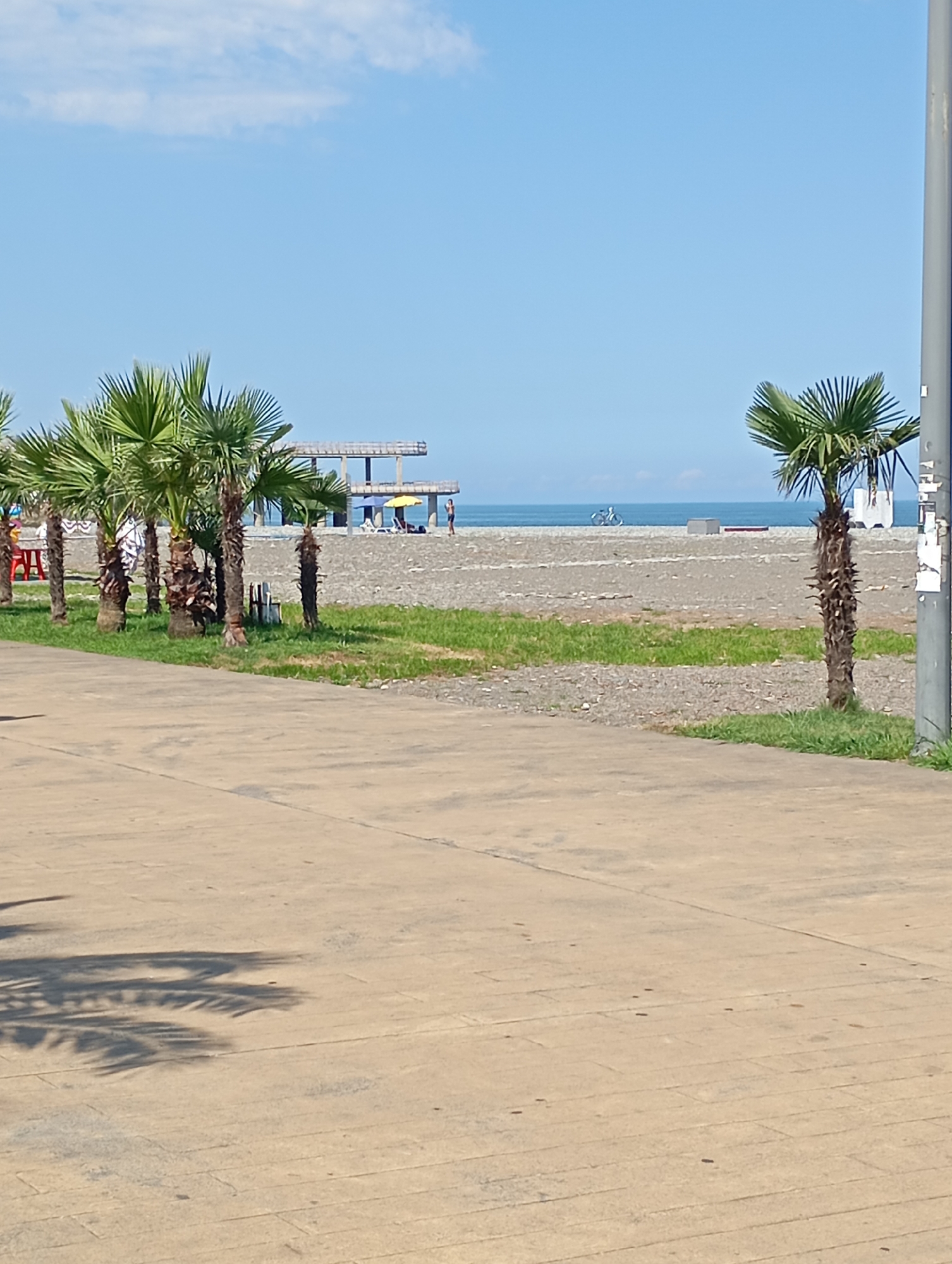 The image depicts a scene at a beach in Batumi, Georgia, under a clear blue sky with scattered white clouds. In the foreground, a light-colored paved walkway with shadows of palm trees is visible. Beyond the walkway, a strip of green grass separates it from a pebble beach. Several palm trees are planted along the edge of the grass, some with mature, textured trunks.
The pebble beach stretches towards the sea, with a few people scattered across it. Some are near lounge chairs and a yellow umbrella, suggesting a relaxation area. In the background, the sea meets the horizon, and a structure resembling an unfinished building or pier is visible. A bicycle is also present on the beach, near the water's edge. A tall, grey pole stands on the right side of the image, partially obscuring the view. The overall impression is a sunny day at a coastal resort.