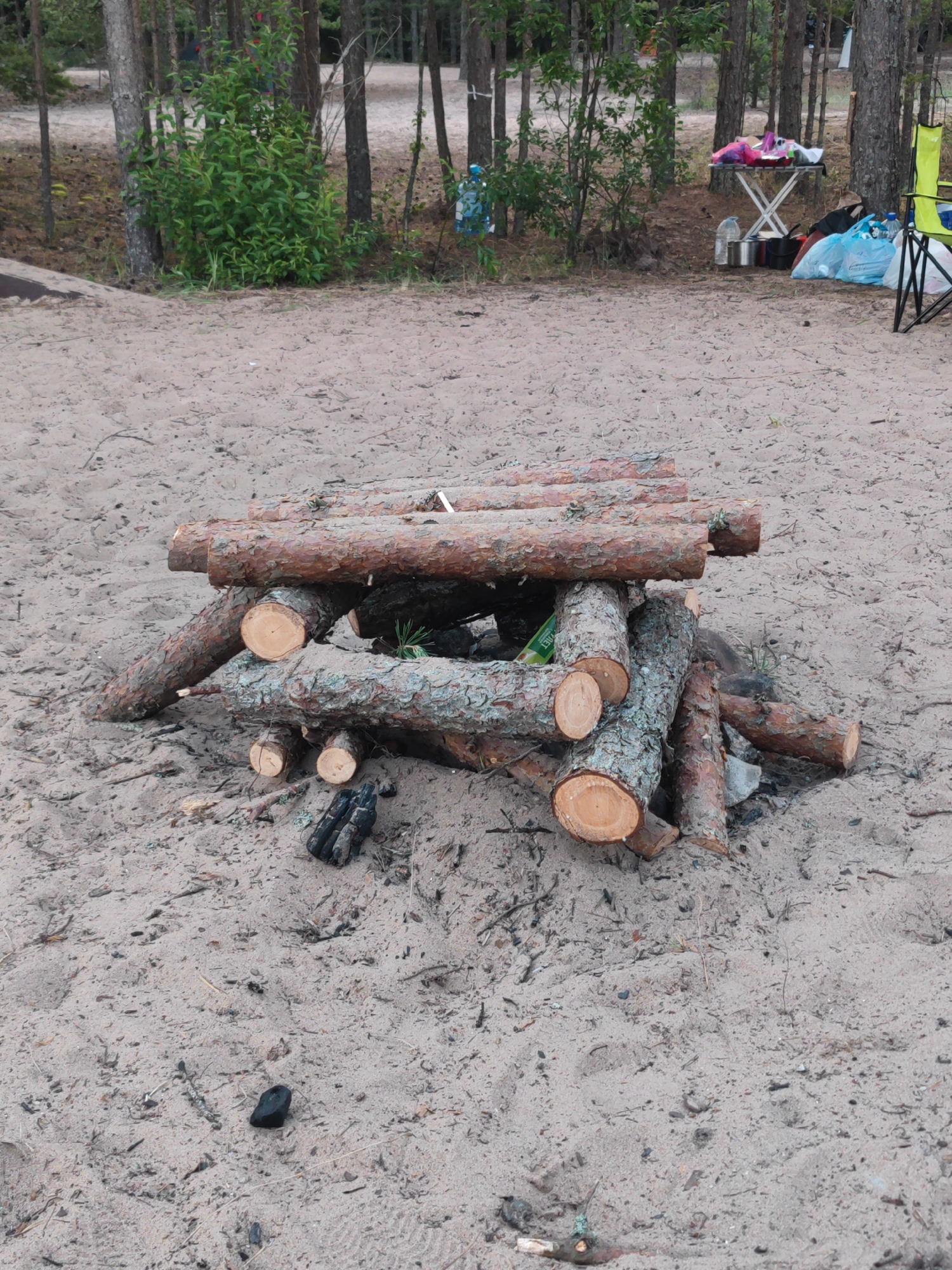 The image depicts a sandy clearing in a pine forest, likely in the vicinity of Zaostrov'e, Russia, given the vegetation and general landscape. In the foreground, a pyre of cut pine logs is arranged, some cross-hatched and others leaning against the base. The logs vary in diameter and show visible bark and wood grain. Scattered around the pyre are remnants of burnt wood and charcoal. In the background, the forest consists of mature pine trees with a sparse undergrowth of green shrubs. Further back, a camping setup is visible, including a foldable table laden with items like bottles, bags, and what appear to be food containers. A bright yellow camping chair is also present. The ground is covered in fine, light-colored sand with scattered pine needles and debris. The overall scene suggests a temporary campsite in a natural environment.