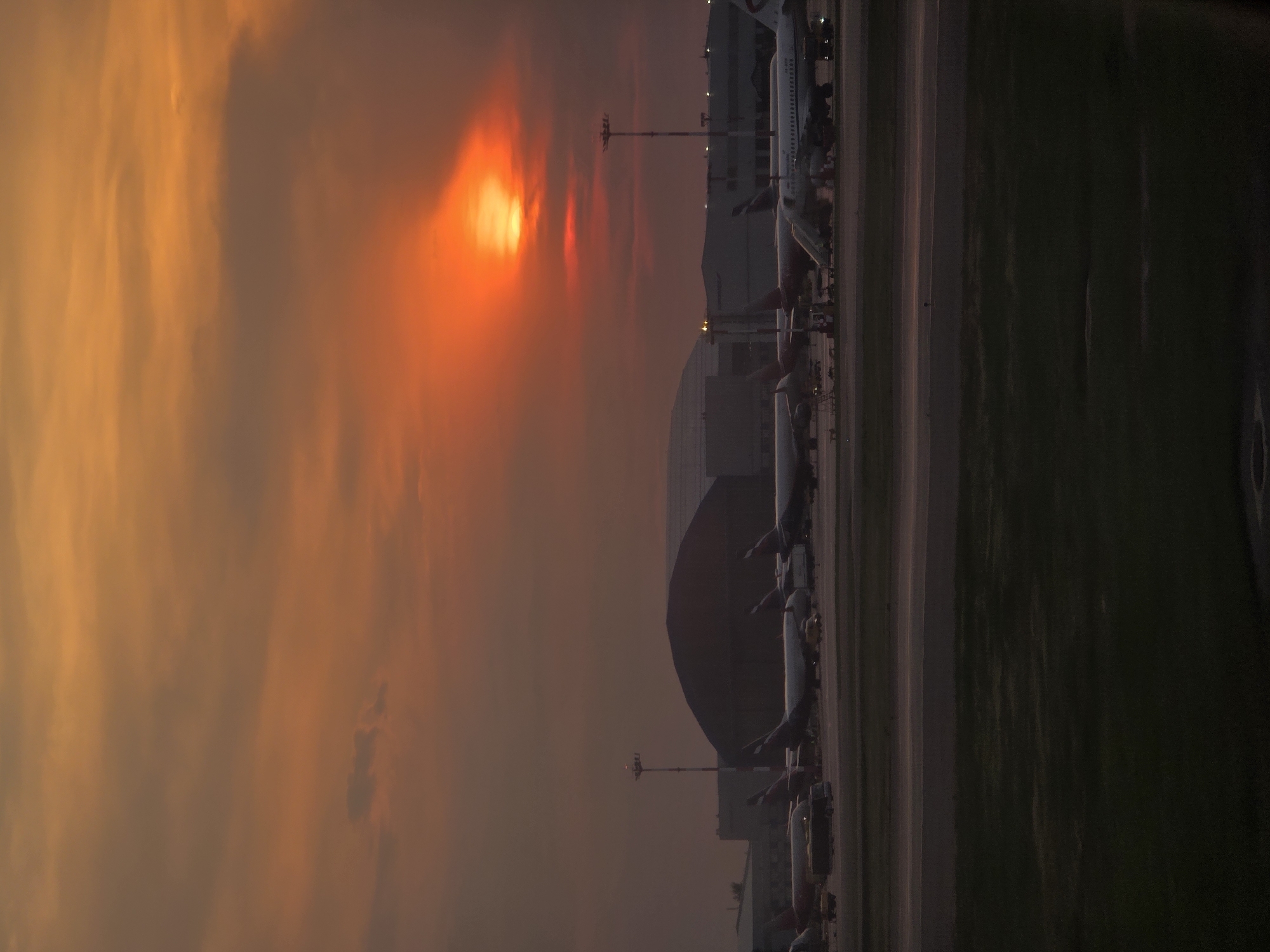 The image depicts an airport tarmac at sunset, with the sun casting a dramatic orange glow across the sky. Several airplanes are parked on the tarmac, some with boarding stairs attached. In the background, large hangar-like structures are visible. The foreground shows a grassy area. The scene is identified as Sheremetyevo International Airport in Khimki, Russia. There are no people clearly visible in the image, and no discernable actions or interactions taking place. The overall mood is serene and atmospheric, characterized by the warm hues of the setting sun.