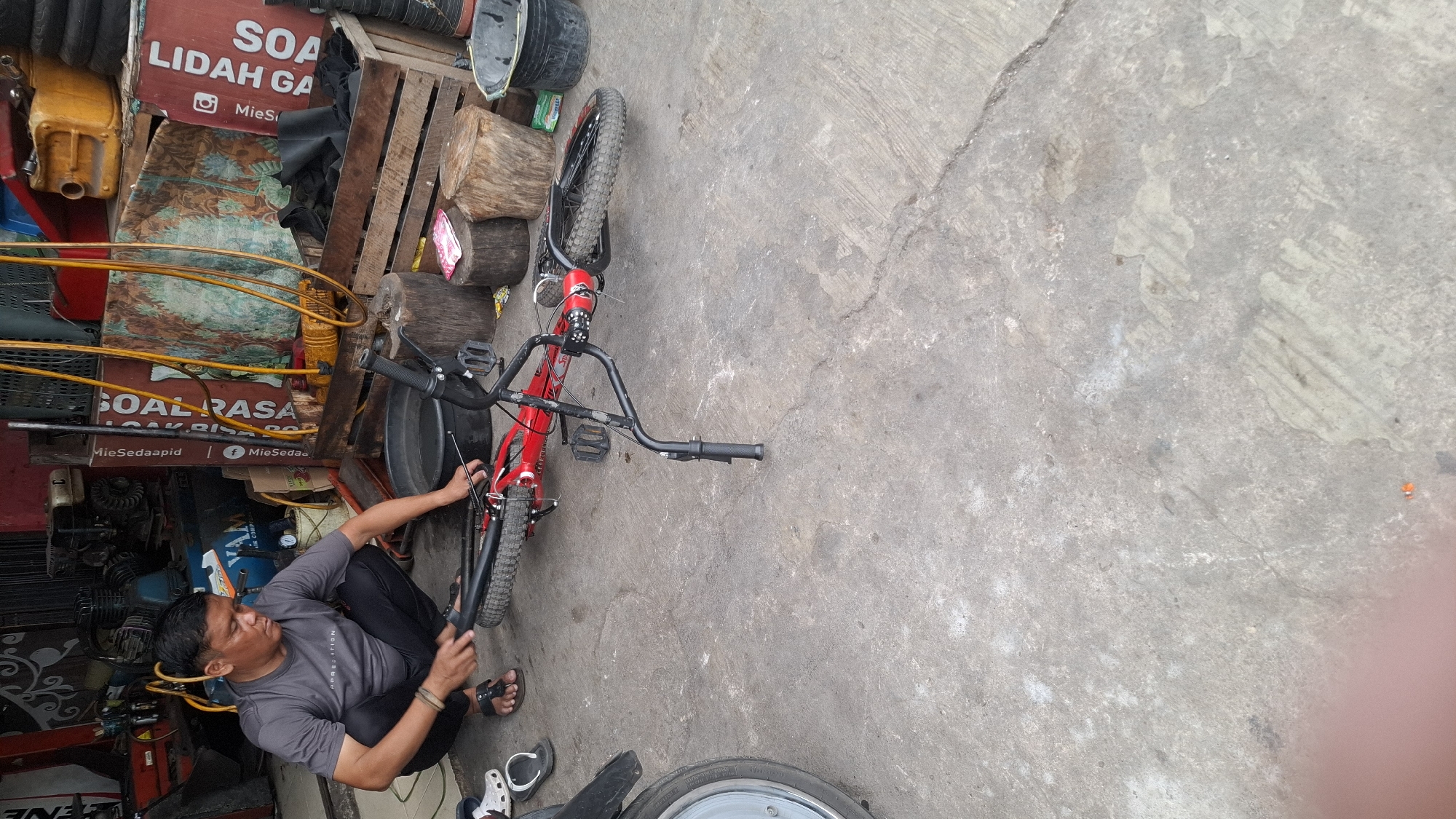 A man is squatting on a concrete floor in what appears to be a workshop or repair shop. He is wearing a gray t-shirt and dark pants. He is working on a red bicycle that is lying on its side. The bicycle has black tires and handlebars. In the background, there are various objects, including a generator, several wooden crates, buckets, and signs. The overall environment suggests a cluttered but functional workspace. The presence of tools and repair equipment indicates that this is likely a place where bicycles are serviced or sold.