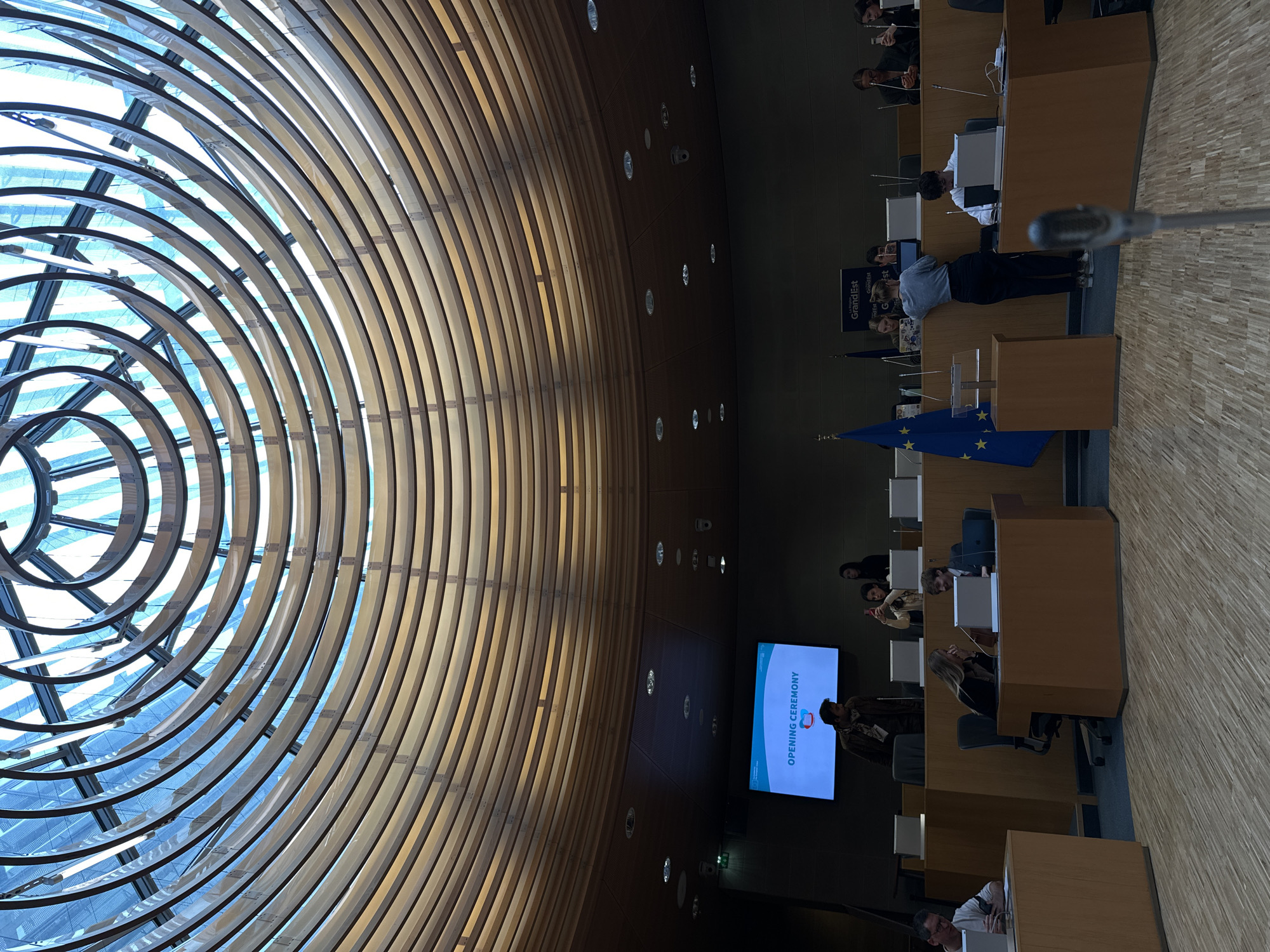 The image shows the European Parliament in Strasbourg, France, during what appears to be an opening ceremony, as indicated by the screen in the background. The room is characterized by a distinctive architectural feature: a large, circular, skeletal-like structure forming the ceiling, which allows natural light to enter the space.
In the foreground, rows of desks are occupied by individuals, some of whom are seated, while others are standing and speaking at podiums. A European Union flag is visible. The environment seems formal, suggesting an official event.