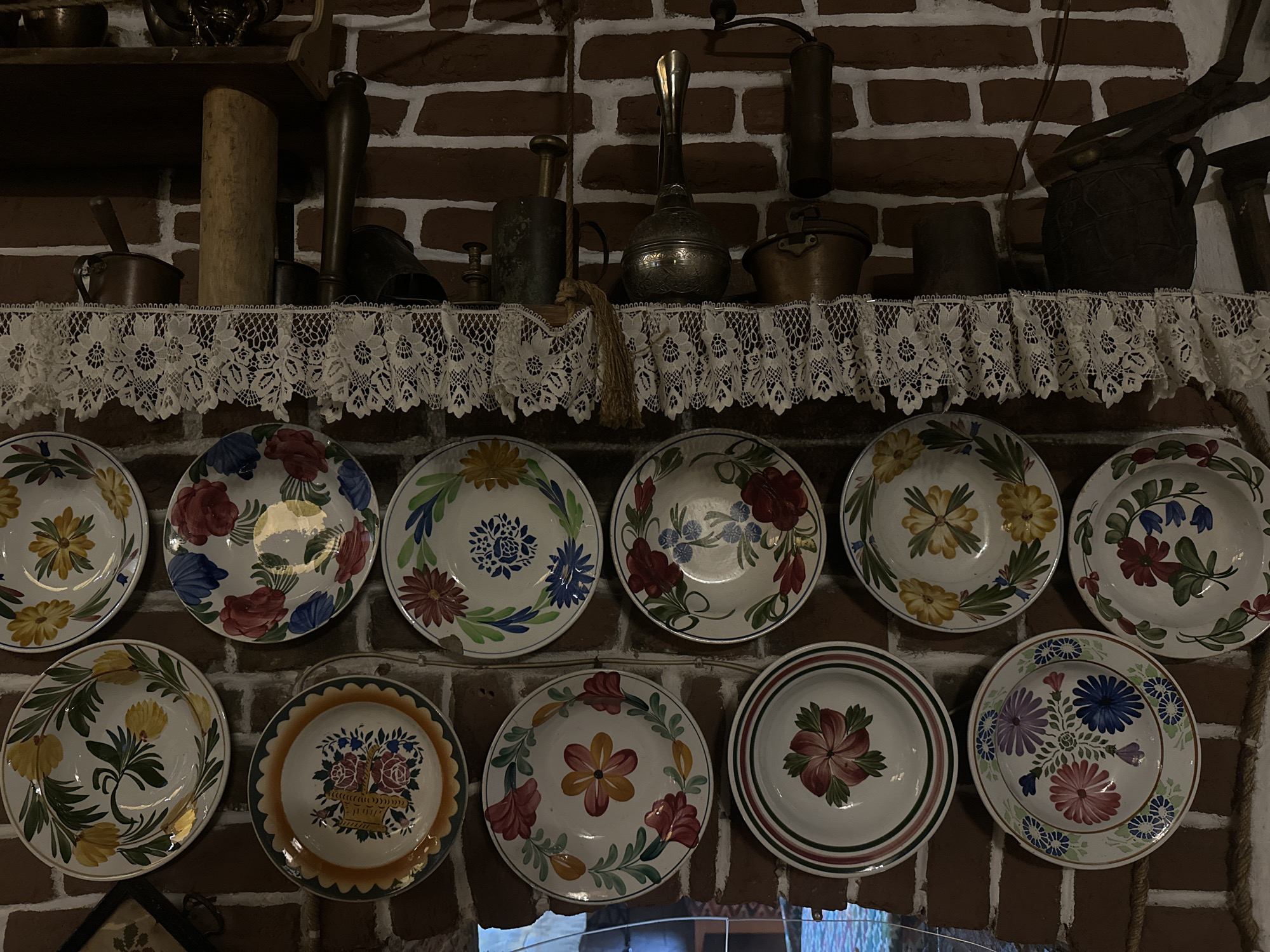 The image shows a collection of decorative plates and antique kitchenware displayed on a brick wall above a lace-trimmed shelf. The plates, adorned with floral patterns and colorful designs, are hung in two rows. Above the shelf, various metal and wooden objects, possibly from a kitchen or workshop, are visible. The overall setting suggests a rustic or vintage aesthetic. The location of this scene is not discernible from the image.