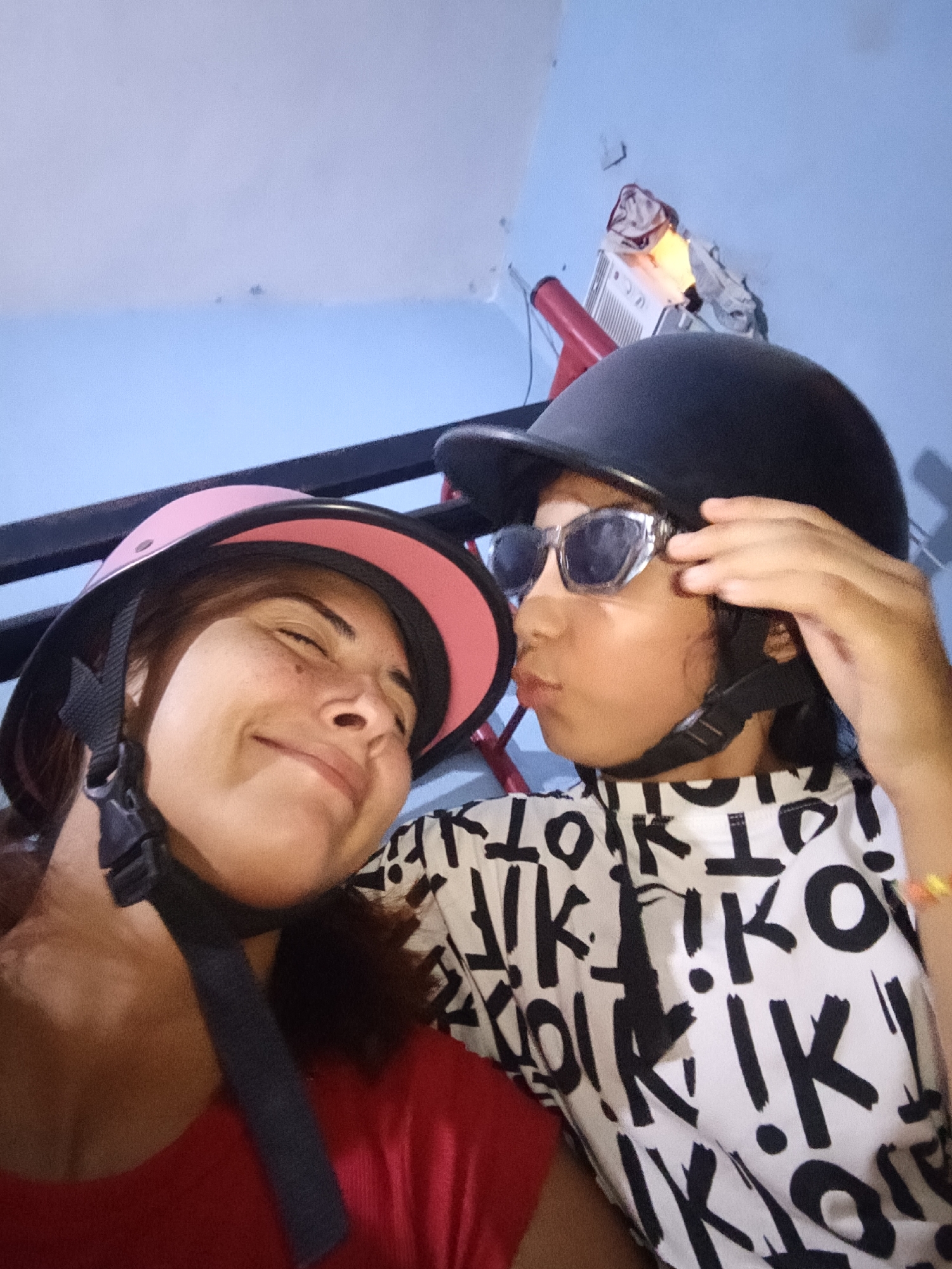 A close-up shot of two individuals in Juan Griego, Venezuela wearing helmets. One individual, presumably a woman, wears a pink-trimmed helmet and a red shirt. She closes her eyes and smiles. The second individual, presumably a child, is in a black helmet and sunglasses, making a kissing face. The child wears a white shirt with black lettering. The location appears to be indoors. In the background, there is a red pole and an air conditioning unit with clothes hanging nearby. The walls are light blue.