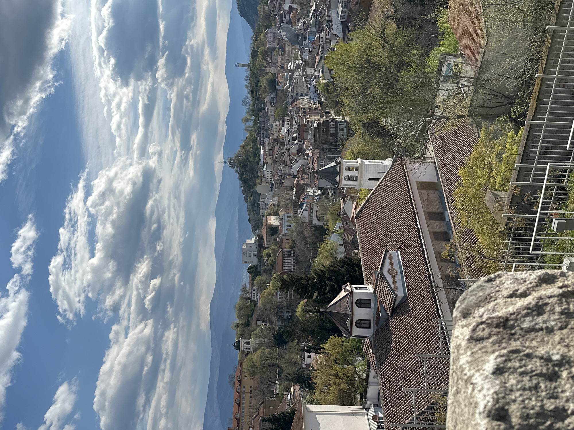 The image shows a cityscape view in Plovdiv, Bulgaria, taken from an elevated position. A blue sky with scattered white clouds occupies the left side of the frame. Mountains are visible in the background, their peaks partially obscured by haze. The city stretches across the middle ground, with numerous buildings featuring red-tiled roofs and various architectural styles, interspersed with trees. In the foreground, there is a stone structure with a textured surface on the right, and a section of a building with a red-tiled roof and a small turret-like structure extending upwards. A metal railing is visible in the right foreground. The image appears to be taken during daylight hours, with the sun casting shadows on the buildings.