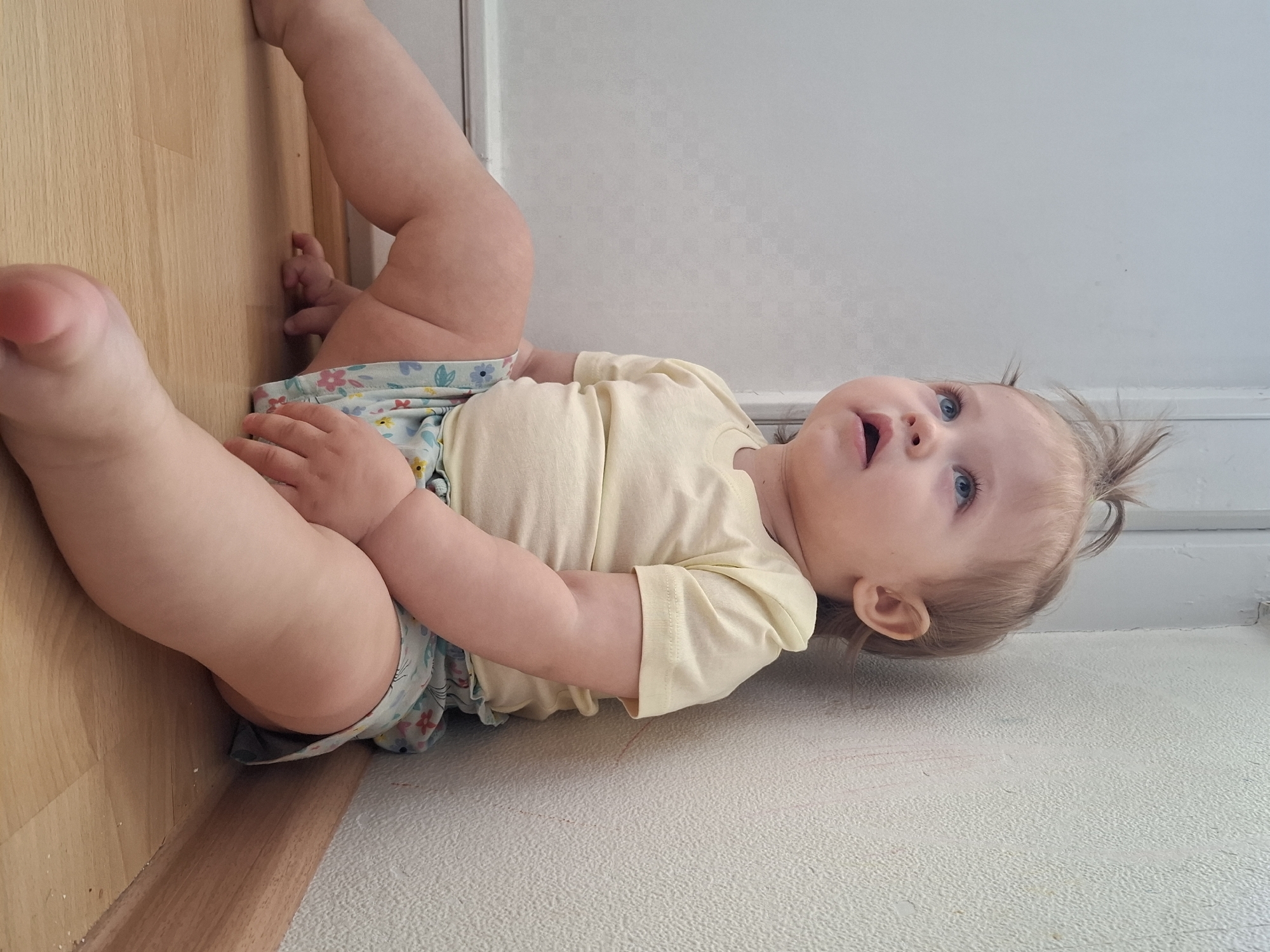 A baby is lying on its back on a textured, white surface, potentially a carpet or rug. The baby's head is near a white wall. To the left, a light-colored wooden floor meets the wall. The baby is wearing a yellow shirt and a diaper with a floral pattern. The baby's legs are raised with one foot near the wooden floor. The baby is looking upwards with an open mouth. It is not possible to confirm the location as Chaikovsky, Russia with the information provided.