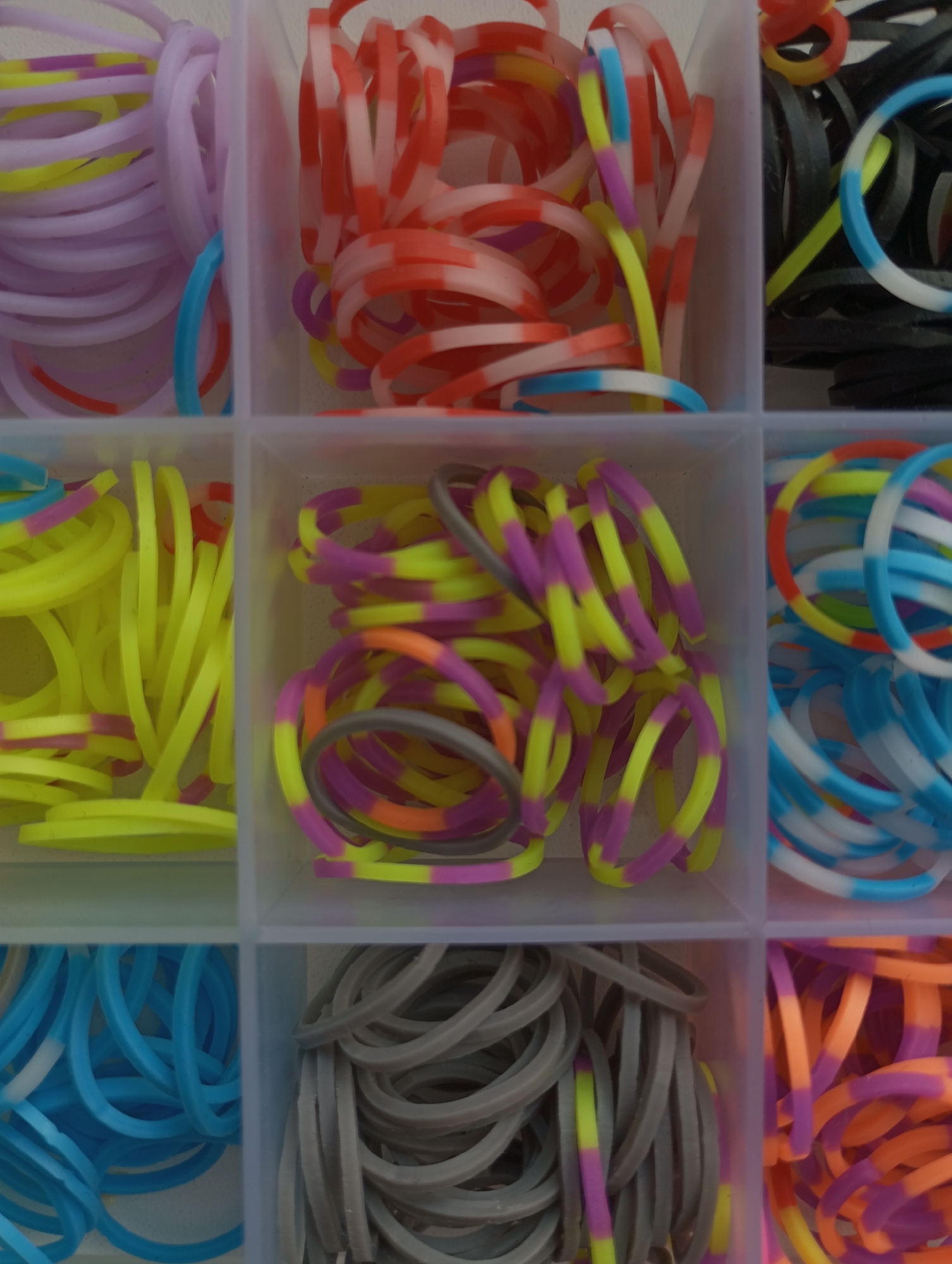 The image displays a close-up, top-down view of a clear plastic organizer box filled with colorful rubber bands, commonly known as loom bands, divided into nine square compartments.
In the top row, the left compartment contains light purple bands mixed with some yellow and pink striped ones, and a loose light blue band. The middle compartment is filled with red and white striped bands, interspersed with yellow, pink, and light blue striped bands. The right compartment holds black bands alongside yellow and light blue/white striped bands.
The middle row features bright yellow bands, some with pink stripes, in the left compartment. The central compartment showcases a vibrant mix of yellow and purple striped bands, with some orange and grey striped bands. The right compartment contains light blue and white striped bands, mixed with orange and red striped ones.
The bottom row includes solid light blue bands, some with white sections, in the left compartment. The middle compartment holds grey bands, some featuring yellow and pink stripes. The rightmost compartment is filled with orange and purple striped bands, with occasional yellow accents.
The rubber bands are neatly organized by color or pattern within their respective compartments, suggesting they are stored for crafting or play. The lighting is even, indicative of an indoor setting. No specific activity is taking place, and no text is visible.