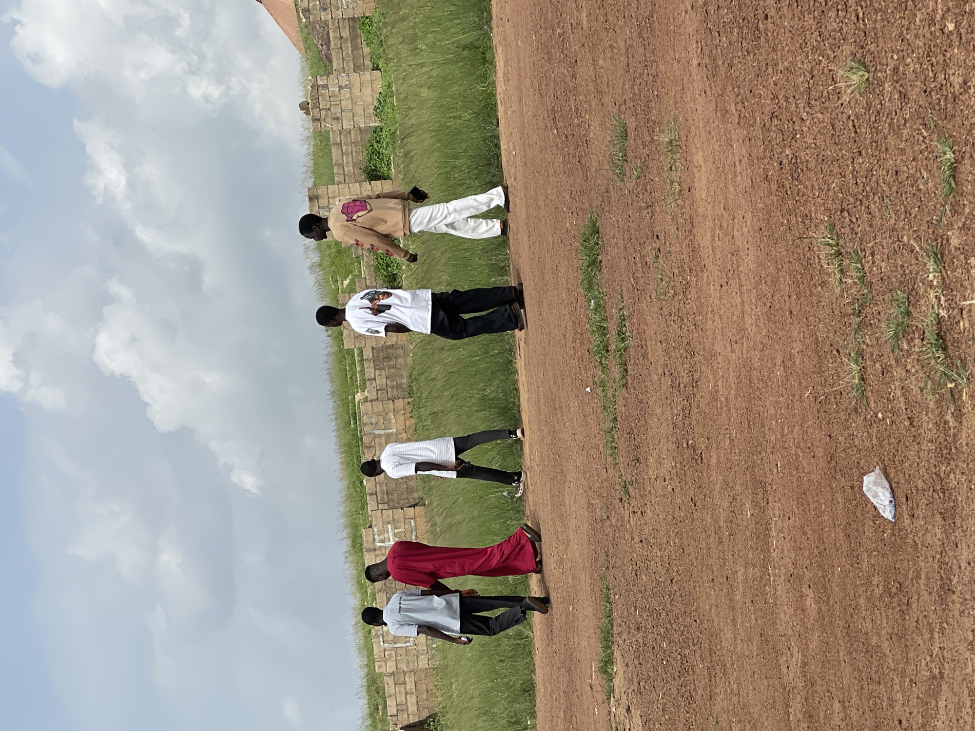 This image, taken in Durumi, Nigeria, shows five young men standing outdoors. They are arranged in a line, facing away from the camera, with the perspective suggesting they are walking towards the viewer. The setting appears to be a dusty, reddish-brown path beside a grassy area with some low-lying brick structures in the background. The sky is partly cloudy, indicating it is daytime.
The men are dressed casually. The person furthest from the camera is wearing a light brown long-sleeved shirt with a graphic design on the back and white pants. Next to him is a person in a white t-shirt and black pants. Following them is someone in a white long-sleeved shirt and black pants. The fourth person is wearing a vibrant red robe-like garment. The person closest to the camera is in a grey t-shirt and black pants.
There is no discernible activity or event taking place, as the individuals are simply standing or walking. The lighting suggests it could be late morning or afternoon. The overall mood is relaxed and casual. A crumpled white plastic bag lies on the dirt path in the foreground.