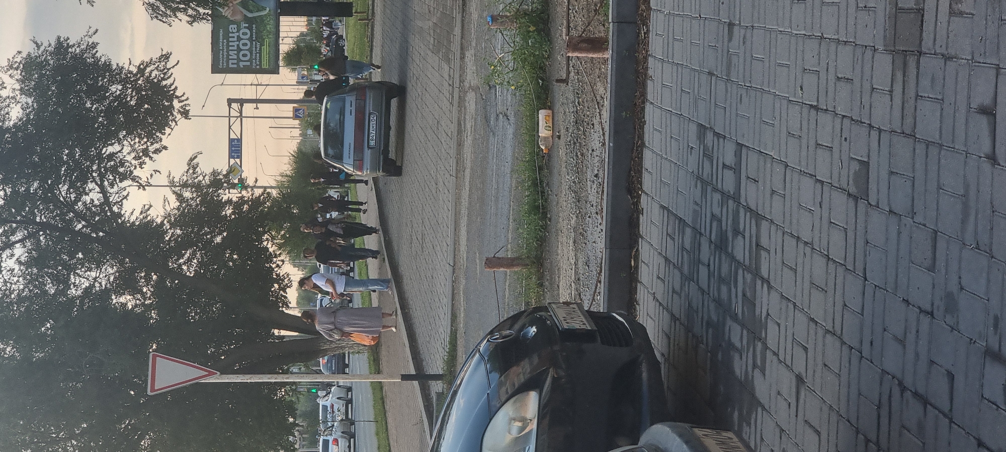 In Aktobe, Kazakhstan, an outdoor scene captures a moment on a sidewalk next to a street. A group of people are walking on the paved sidewalk. A woman in a long dress and carrying an orange bag walks with a man in a white t-shirt and jeans, and another woman in black. Behind them, a group of men and women walk in the same direction. A silver car is parked on the street. In the foreground, the front of a black car is visible. A yield sign is mounted on a pole near a large tree. In the background, a billboard advertises pizza. The sky is overcast, suggesting late afternoon or early evening.