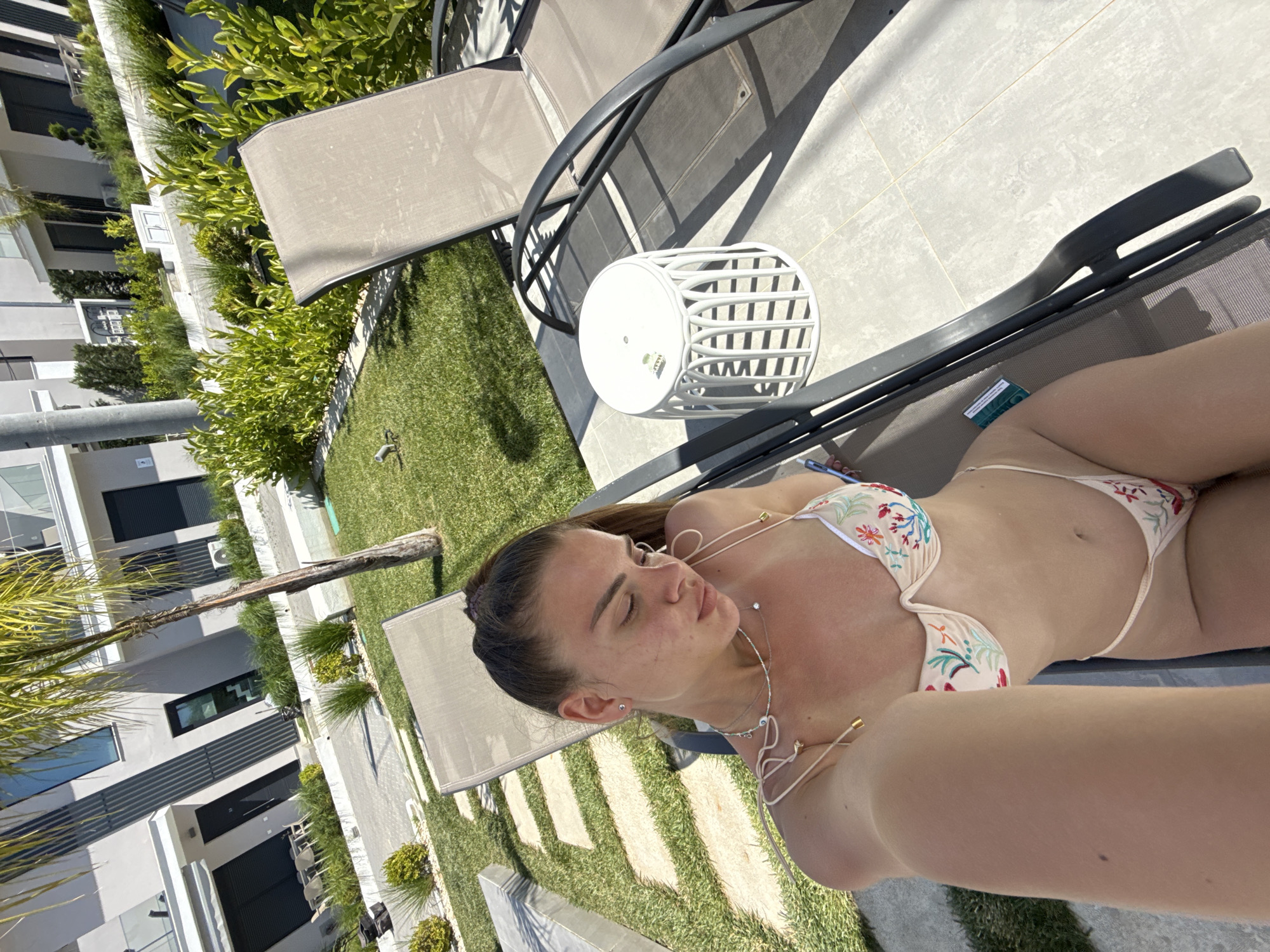 A daytime selfie shows a woman reclining on a deck chair outdoors. She is wearing a floral bikini and several necklaces, and her eyes are closed. A white, cylindrical stool sits next to the deck chair. The surrounding environment includes green grass, a tiled patio, and several buildings, possibly apartments or condominiums.