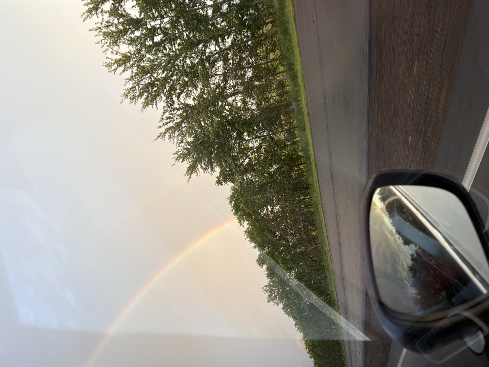 The image captures a view from inside a vehicle, looking towards the side mirror and out the window. A faint rainbow arcs across the pale sky on the left side of the frame. A line of trees, appearing dense and green, runs parallel to a dark asphalt road. The road has white lane markings and appears to be wet, possibly from recent rain. Reflections of the sky and trees are visible in the side mirror, which is black and mounted on the vehicle. The overall impression is a travel scene, likely on a highway, with the natural phenomenon of a rainbow present. The location is identified as Таймурзино, Russia.