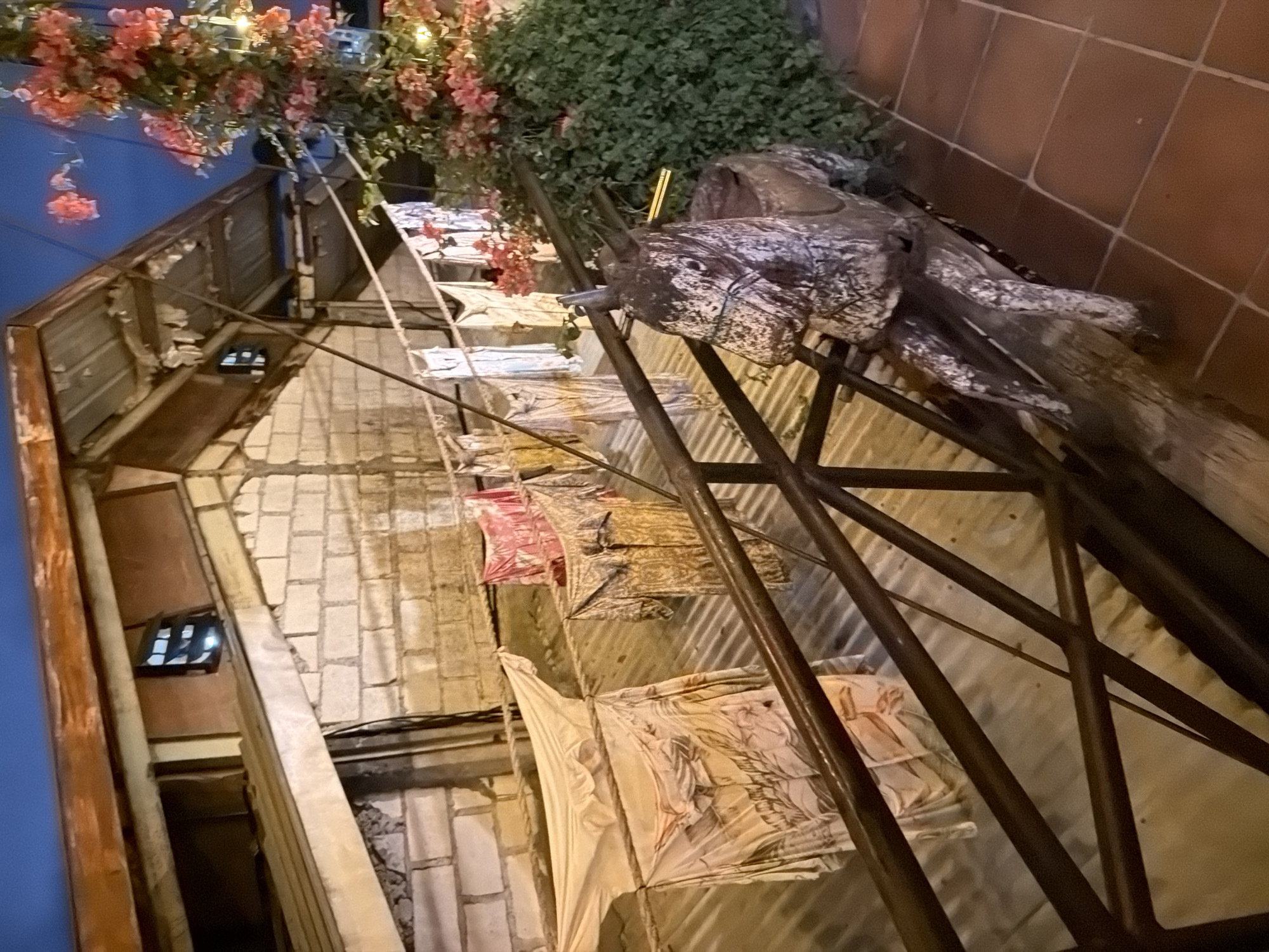The image shows a clothesline with several garments hanging to dry in front of a brick building with a corrugated metal roof. A wooden rocking horse sculpture is positioned on the right side of the frame, on a tiled floor. Pink bougainvillea flowers and green foliage are visible in the background. The scene appears to be outdoors, possibly a residential area or a small business in Kuta, Indonesia. The lighting suggests it is either dusk or nighttime, with artificial lights illuminating the area.