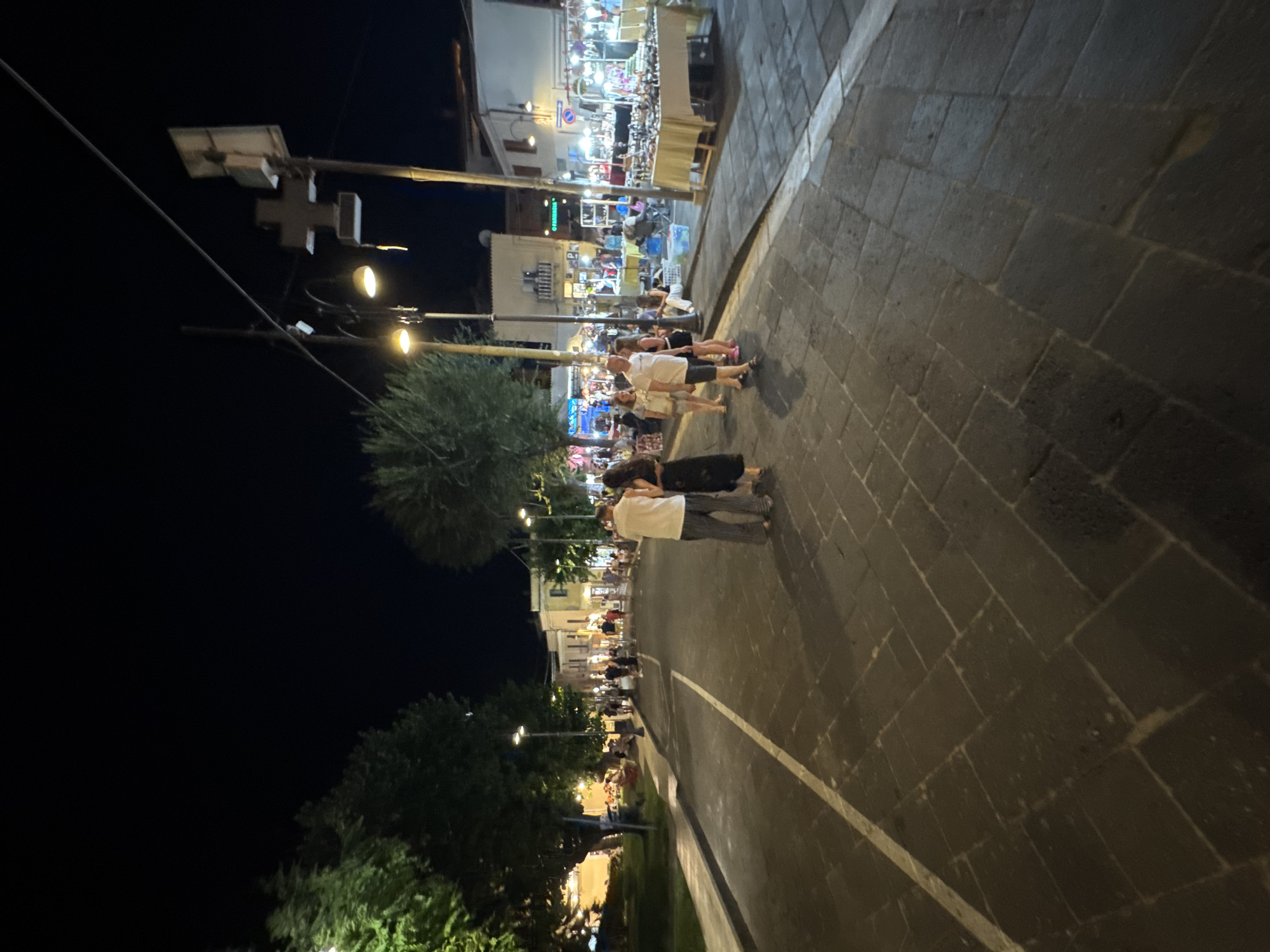The image depicts a street scene at night in Orosei, Italy. A paved street with cobblestone-like sections and a white line marker runs through the center of the frame. On the right side of the street, there are several market stalls displaying goods, illuminated by overhead lights. People are browsing and interacting at these stalls. On the left side of the street, a grassy area with trees is visible, also illuminated by streetlights. Several people are walking along the sidewalk and street. In the foreground, two individuals stand facing away from the camera, with one person wearing a white top and striped pants, and the other wearing a dark dress. The overall atmosphere suggests a lively evening in a town square or main street with shops and activities.