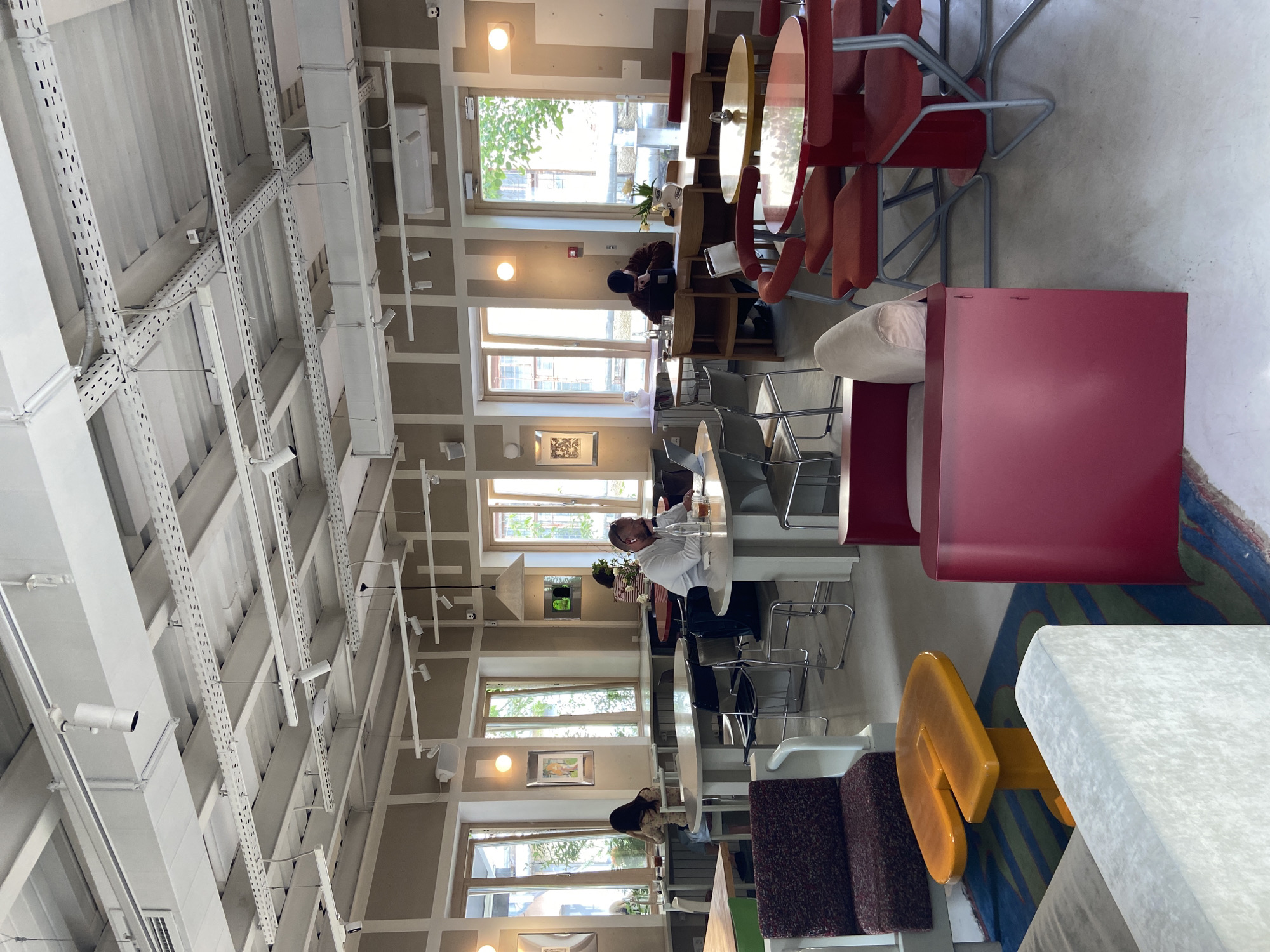 The image depicts the interior of a cafe or co-working space, possibly in Moscow, Russia. The space is characterized by its high ceilings with exposed white structural elements and extensive ductwork. Numerous windows line the walls, providing natural light.
The seating arrangement includes a variety of chairs and tables, including red cantilever chairs and round tables. People are seen seated at some of these tables, working or conversing. One individual is wearing a white shirt and appears to be actively using a laptop. Other seating options include cushioned benches with colorful striped rugs underneath. The environment seems designed for a mix of individual work and casual social interaction. There's an overall modern and minimalist aesthetic. Security cameras are mounted on the ceiling.
