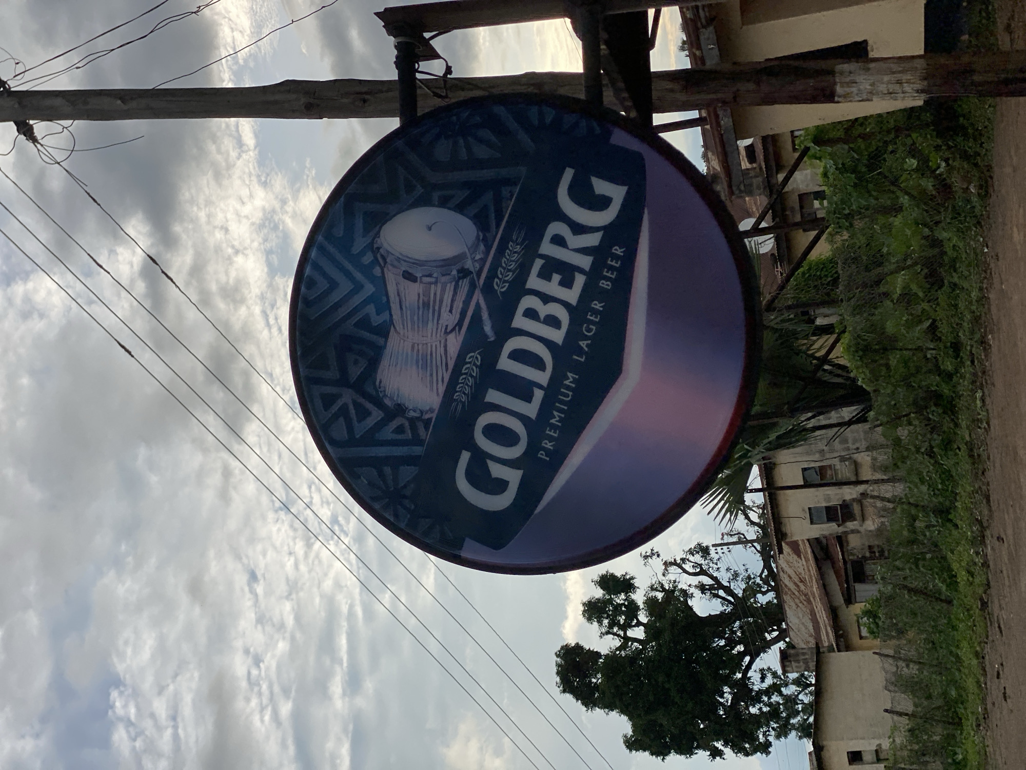 A circular Goldberg Premium Lager Beer sign hangs from a wooden pole next to a street, with power lines strung across the cloudy sky. The sign displays a blue design with images of drums and wheat stalks, and the word "GOLDBERG" in large white letters. Behind the sign, there are several buildings with weathered roofs and overgrown vegetation. The buildings appear to be in a state of disrepair. The overall scene suggests a rural or semi-urban setting in Maigizo, Nigeria.
