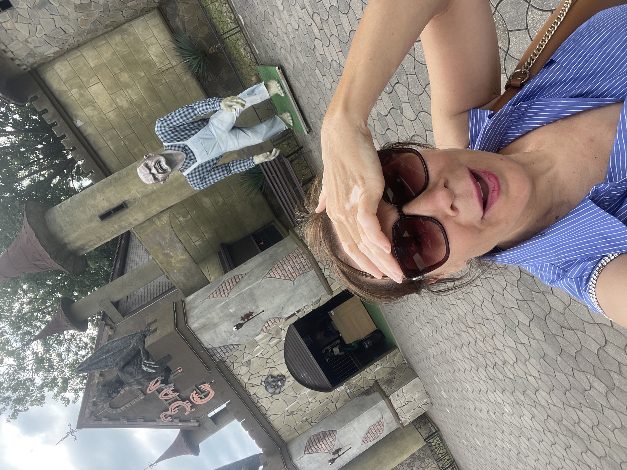 This is a selfie taken by a woman with sunglasses and a blue and white striped shirt. She has her hand on her forehead. Behind her, to the left, is a spooky-themed attraction with a mock castle structure and a scarecrow-like figure in a plaid shirt and overalls. The scarecrow has a grotesque face and outstretched arms. The attraction features stone-like walls and conical towers. There is also some greenery and trees visible. The ground is paved with interlocking stones. The overall mood of the scene appears to be playful and perhaps a little eerie, given the attraction's theme. The lighting suggests it is daytime, and the sky appears overcast. The provided location context is Sochi, Russia, which may imply a tourist or amusement park setting. No visible text is present in the image.