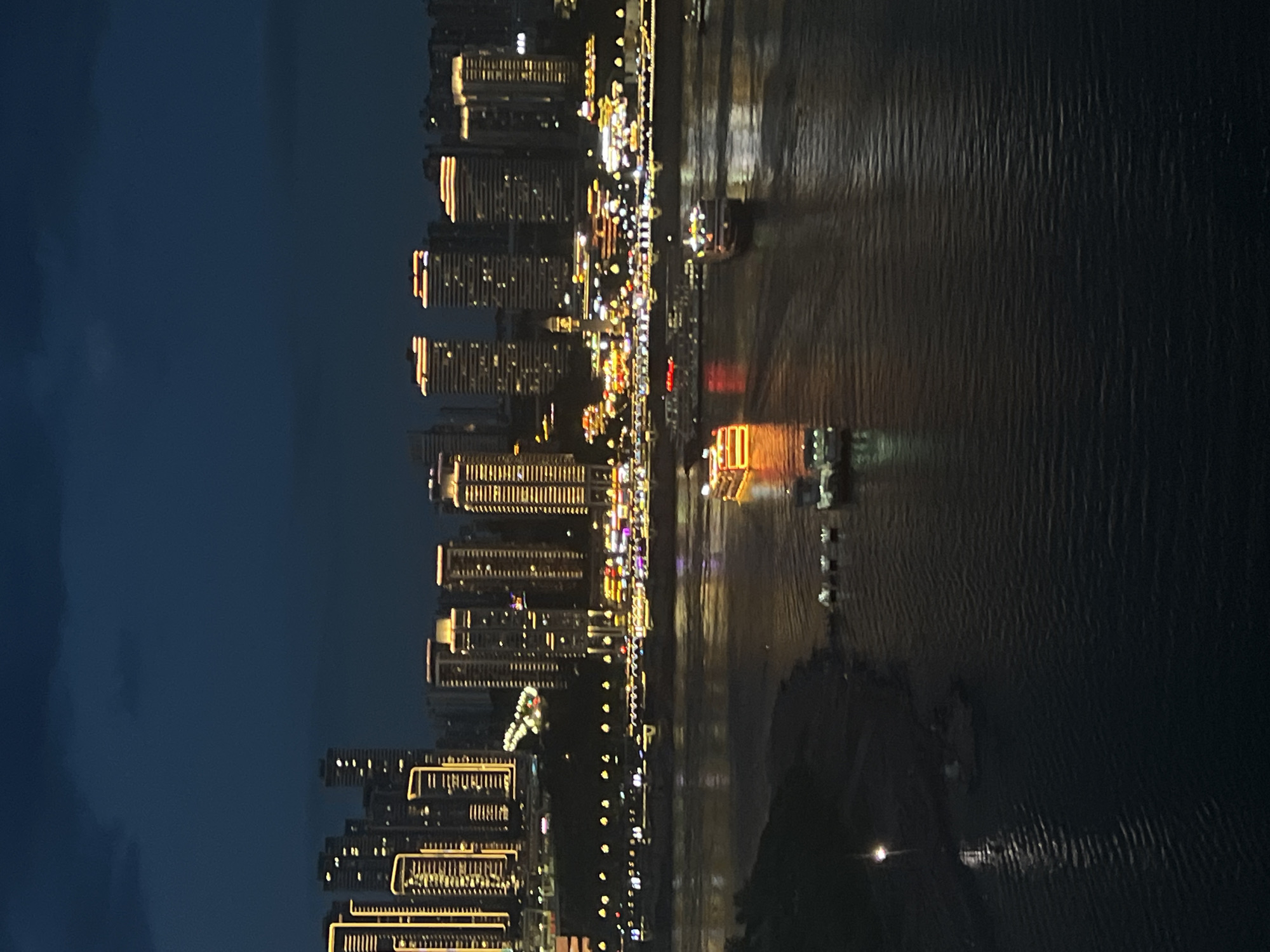 This is a nighttime photograph of a cityscape at dusk, overlooking a wide river. In the foreground, the dark water reflects the lights from the city and from several boats. One prominent boat in the middle of the river is illuminated with bright yellow and orange lights. Further back, smaller boats are visible.
The middle ground features a densely packed cityscape with numerous illuminated skyscrapers and buildings, their windows glowing with yellow light. A waterfront promenade is also visible, lined with more lights. The buildings are of varying heights and architectural styles.
The background is dominated by the dark night sky, with hints of clouds. The overall impression is one of a vibrant and active urban environment at night, likely a port city. The scene is consistent with the Yuzhong District in Chongqing, China, known for its dramatic skyline and location at the confluence of the Yangtze and Jialing rivers.