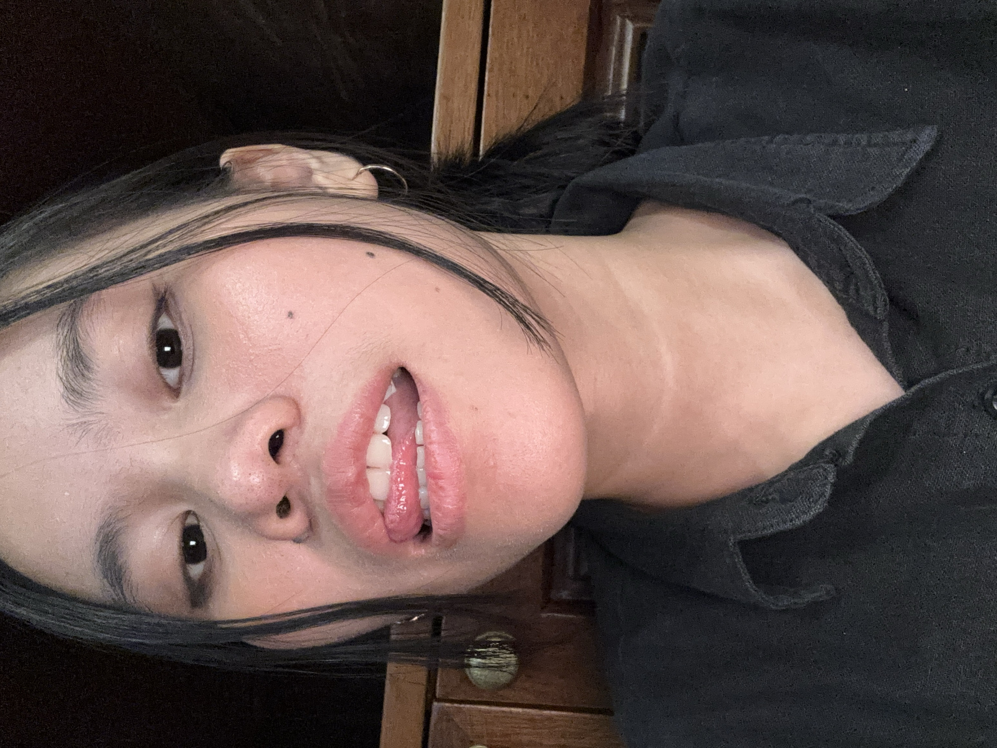 A young woman with dark hair, wearing a black collared shirt, is shown in a close-up shot. She has her tongue sticking out, lightly touching her lips. She has a mole on her right cheek and is wearing a hoop earring in her visible ear. The background appears to be a wooden dresser. The lighting is warm, suggesting an indoor setting. The image was taken in South Jakarta, Indonesia.