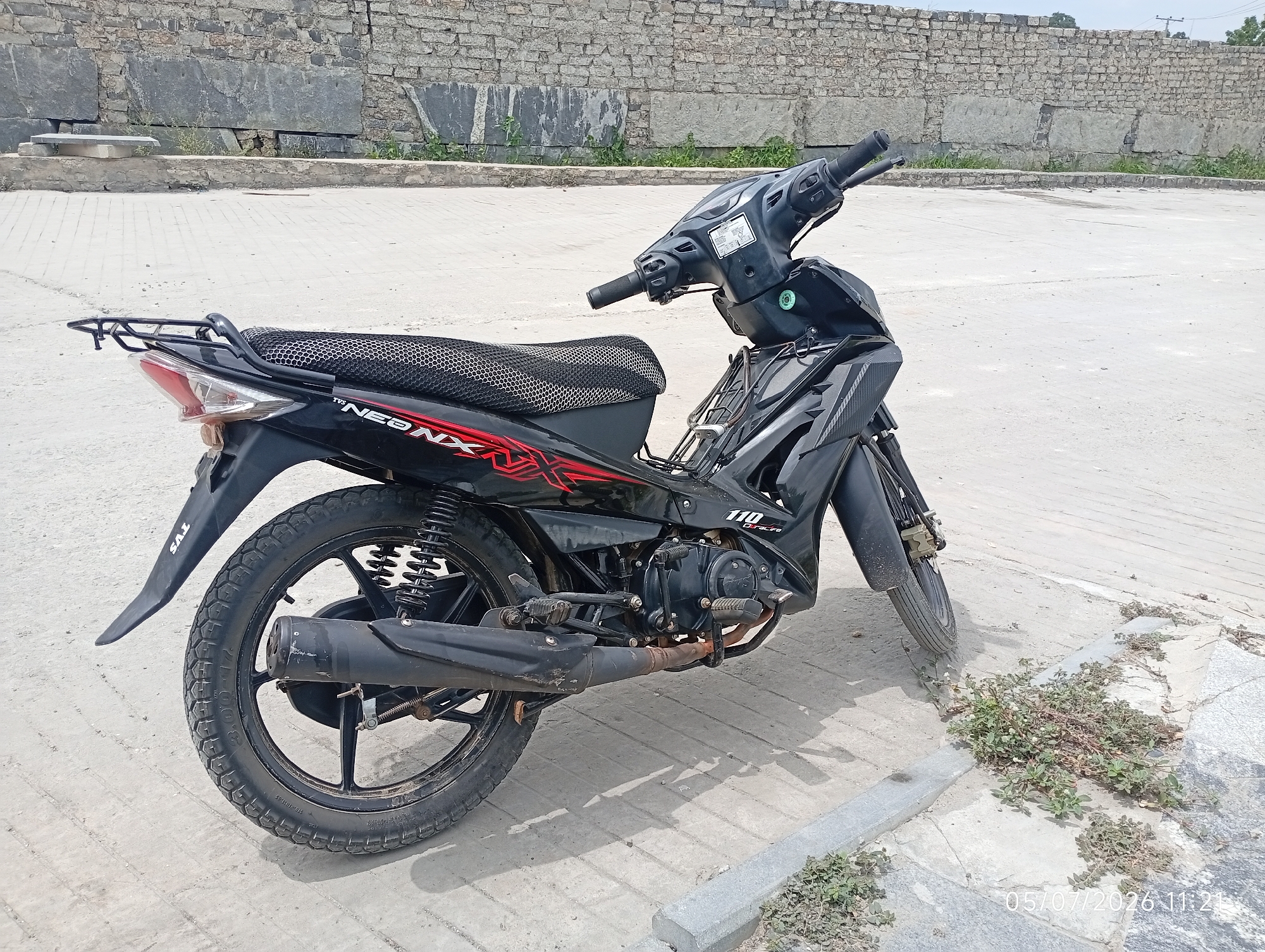 A black TVS Neo NX 110cc motorcycle is parked on a paved surface next to a low stone wall in Bana Stream, Nigeria. The motorcycle is oriented facing the right side of the frame, with its front wheel slightly turned. The scene appears to be outdoors, under daylight, with a clear sky. The background consists of the stone wall and a large, open paved area. Some sparse vegetation is visible growing along the edge of the paved area. The motorcycle shows signs of use, with some dust and wear visible on its components. No people are present in the image.