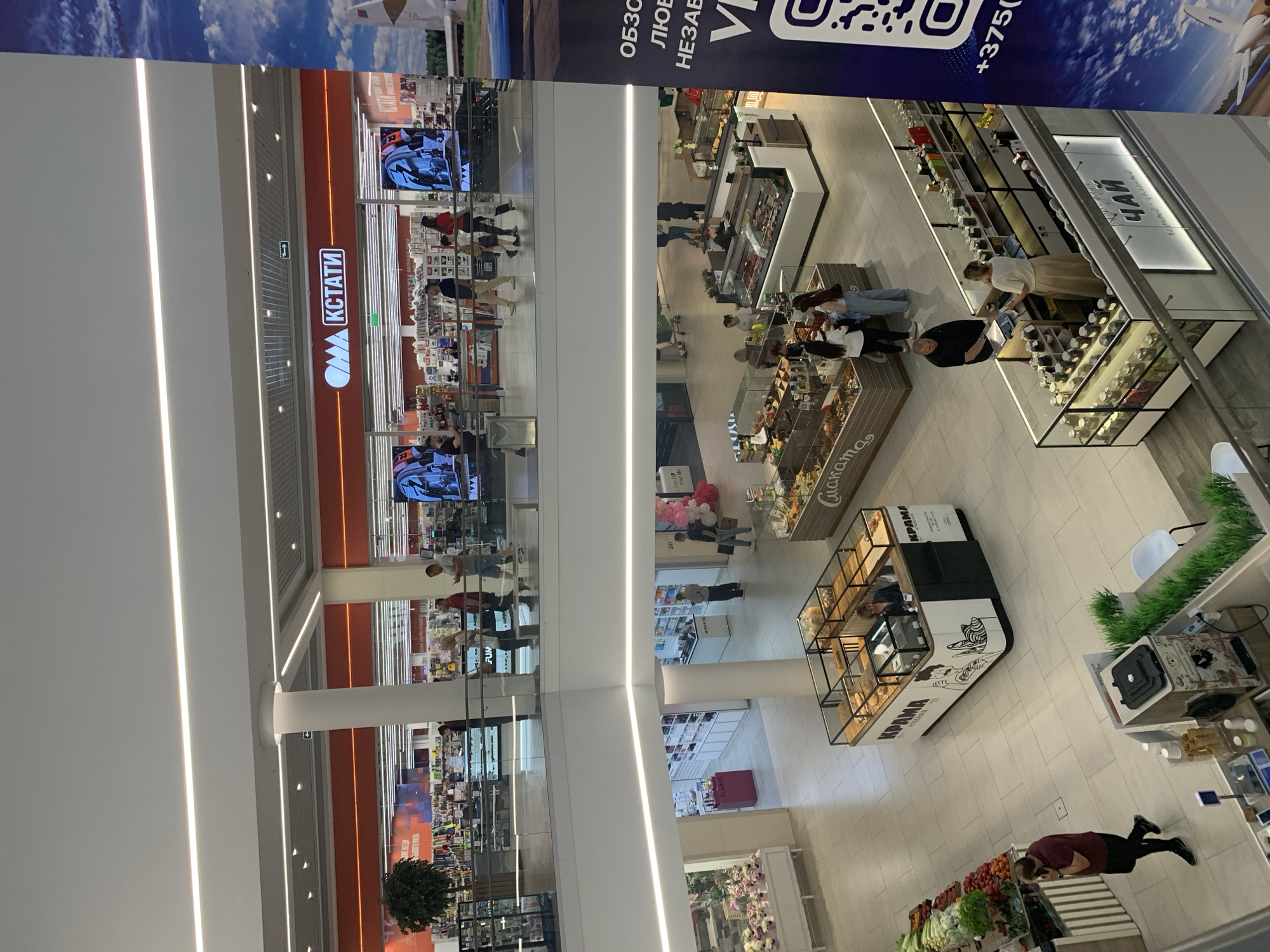 The image is an interior shot of a modern shopping mall, likely located in Hrodna, Belarus, given the Cyrillic signage visible. The perspective is from an upper level looking down onto multiple levels of the mall.
On the upper level, a large orange banner with white lettering reads "КСТАТИ" (Kstati), indicating a store name. Several people are walking along a balcony area in front of this store. Below this level, another section of the mall is visible with various shops. One shop features a prominent display of flowers and plants. Further down, a bakery stall with glass displays showcasing bread and pastries is present, with the Cyrillic word "КРАМА" (Krama) visible on its facade. Adjacent to the bakery is another retail space, possibly selling teas, with shelves stocked with jars and labeled in Cyrillic with "ЧАЙ" (Chai). A man in a black shirt is interacting with a woman at this tea counter. In the foreground of the lower level, a person in a maroon shirt is walking and speaking on a phone, near a display of fresh produce including lettuce, tomatoes, and peppers. Another stall with glass display cases filled with food items is also visible. A large blue banner with text and a QR code is on the far right edge of the image. The mall features bright lighting and modern architecture with white walls and light-colored flooring.