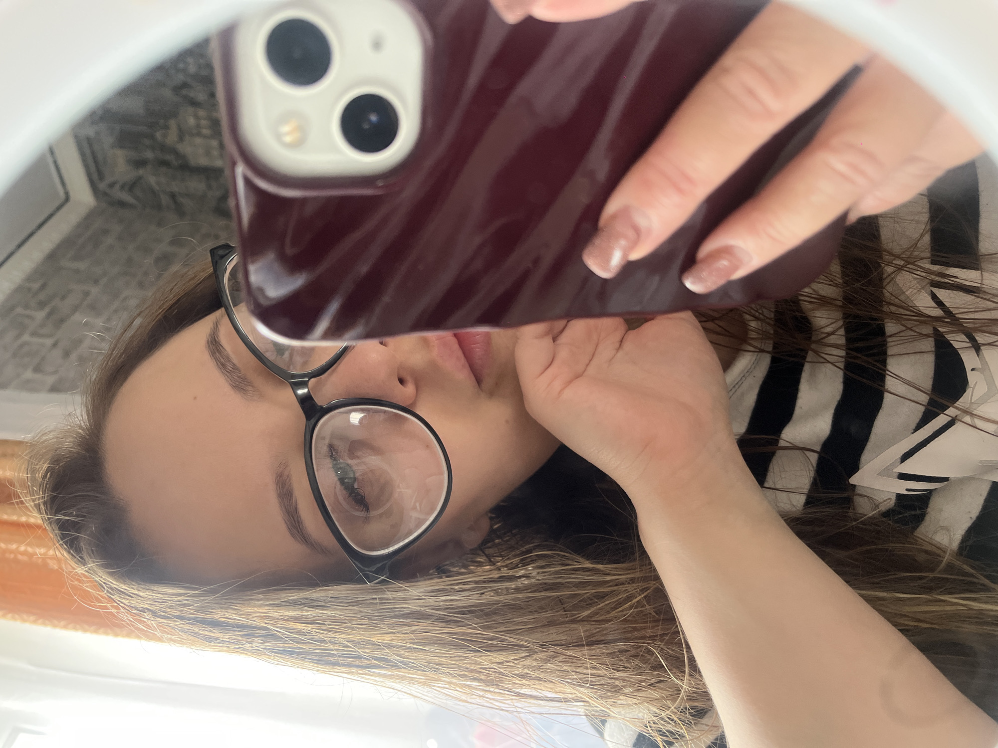 A woman with light brown hair is taking a selfie in a mirror, likely using a ring light. She is wearing glasses with thick black frames and a black and white striped top. Her right hand, showing manicured nails with a nude polish, holds a dark red phone with a triple-camera setup up to her face. The background appears to be an interior space, possibly a home, with a glimpse of a brick-patterned wall visible in the upper left. The location is Novopokrovskaya, Russia.