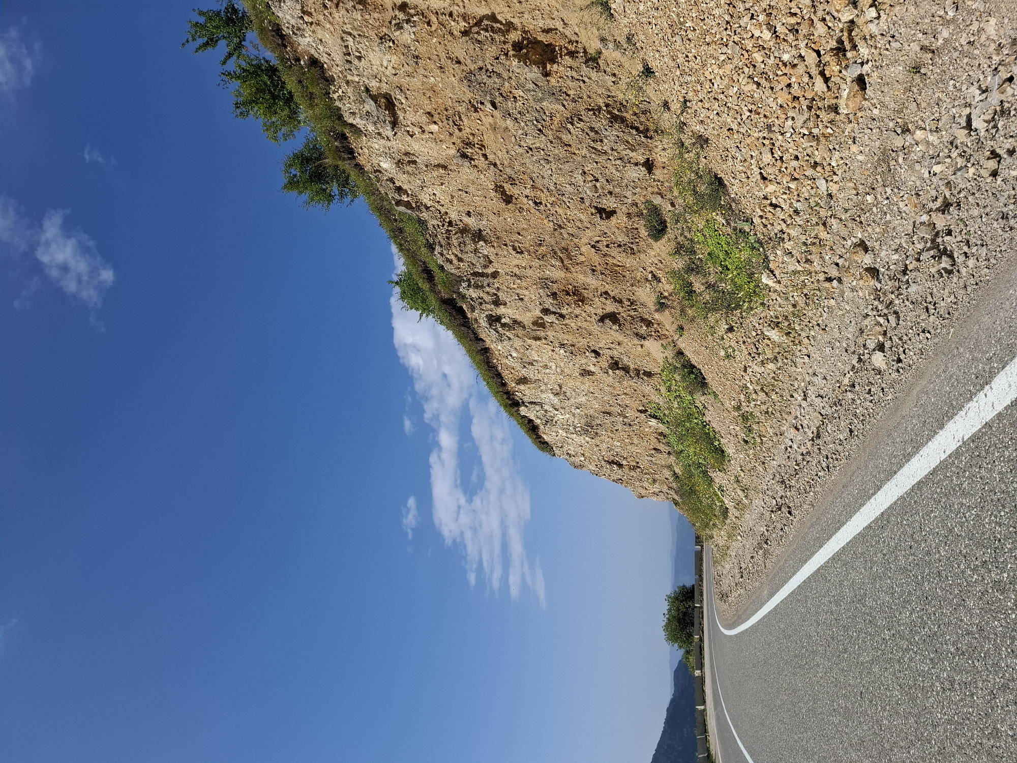 The image depicts a scenic view of a winding asphalt road with a white line marking its edge, curving to the left. To the right of the road, a steep, rocky embankment rises, composed of loose soil and stones in shades of brown and orange. Patches of green vegetation, including small bushes and grass, cling to the embankment. At the top of the embankment, a few trees are visible, their leaves a vibrant green against the clear blue sky. The sky is bright and expansive, with a few scattered white clouds. In the distance, rolling hills with a hazy, muted blue hue suggest a mountainous landscape. The overall scene suggests an outdoor environment, likely a rural or mountainous region, given the presence of the road and natural terrain. The photograph does not contain any people or animals, nor does it show any specific buildings or urban structures. The provided crops focus on different aspects of the scene, such as the sky, the embankment, and the road, allowing for a closer examination of these elements. The location is identified as the city of Kharachoi, Russia.