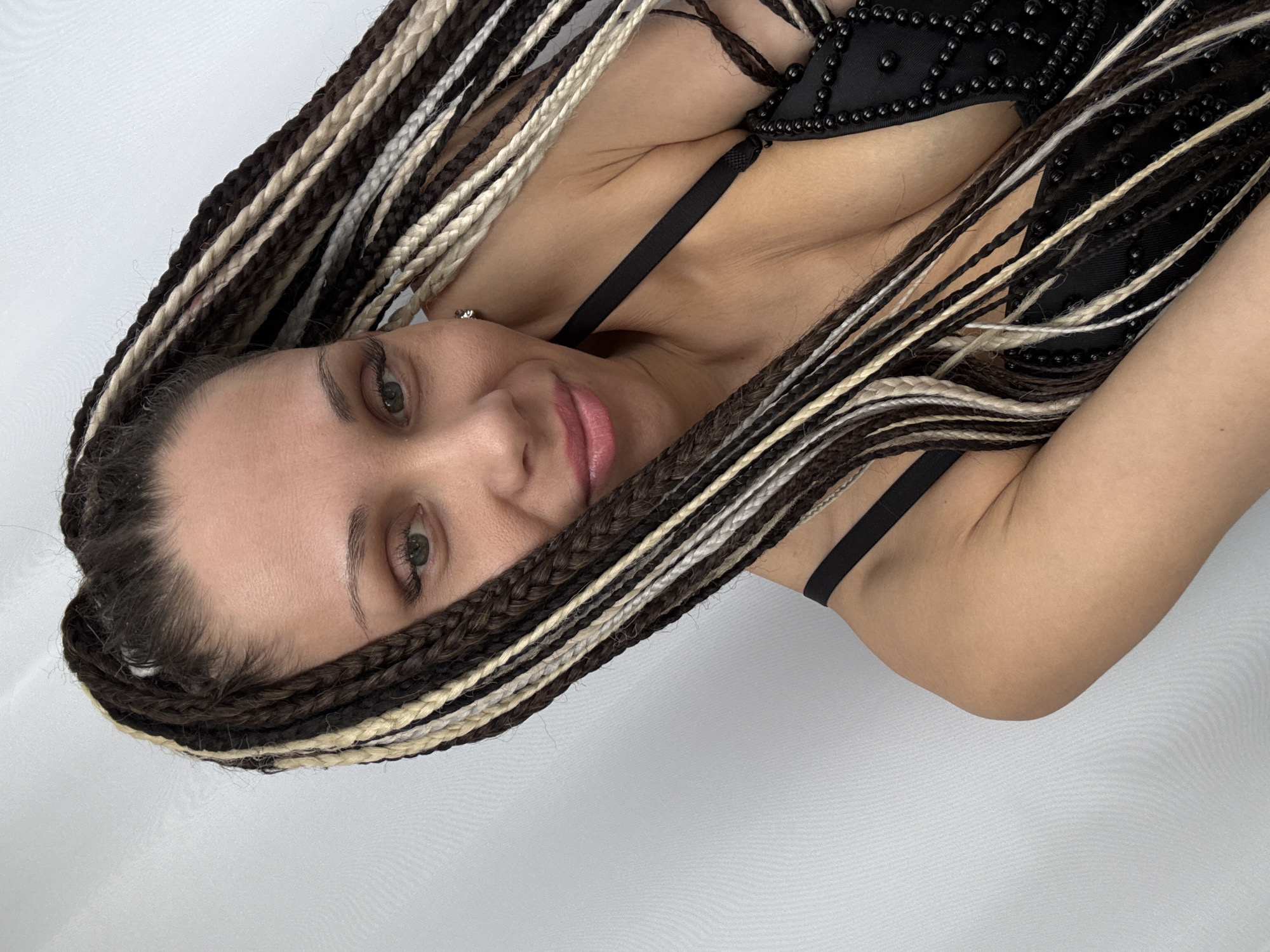 The image shows a woman with braided hair in shades of brown and blonde. She is wearing a black top with beaded embellishments. Her skin tone appears to be fair. The background is a plain white color.