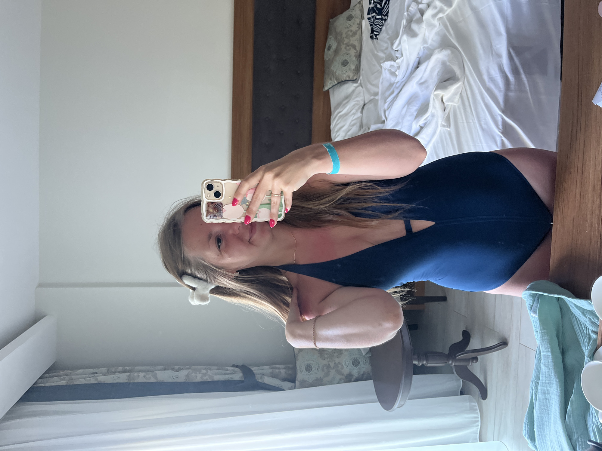 A woman is taking a mirror selfie in what appears to be a hotel room in Alanya, Turkey. She is wearing a dark blue one-piece swimsuit with a deep V-neckline. Her hair is long and brown, with a white fuzzy hair clip on one side. She is holding up her phone, which has a floral patterned case, to take the picture. Her fingernails are painted bright pink, and she is wearing a thin gold necklace and a rose gold bracelet. A bright blue wristband is visible on her left forearm. The room is furnished with a bed with white and patterned bedding, a wooden headboard, a small round wooden table with carved legs, and floor-to-ceiling white curtains. A light blue cloth is draped over a surface in the foreground. The overall impression is of a vacation setting.