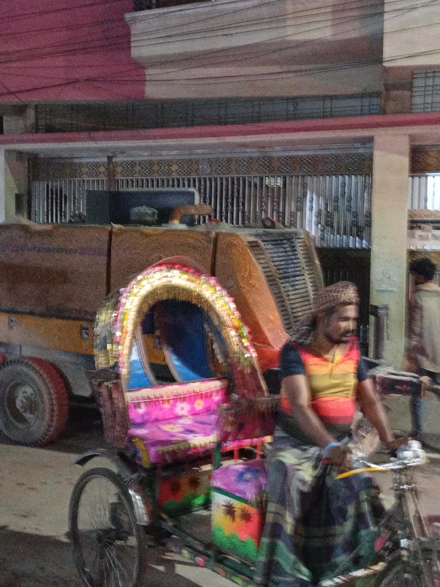 A vertical image captures a vibrant street scene at night, centered around a man operating a brightly decorated rickshaw. The rickshaw, positioned in the foreground, features a prominent pink passenger cabin adorned with multi-colored floral patterns and a canopy embellished with shiny, multi-colored tinsel or lights. Its seat is blue, and one wheel is visible on the left.
The rickshaw puller, a man with a beard and a patterned head covering, wears a striped shirt in orange, yellow, and black, along with a patterned cloth wrapped around his lower body. He is actively engaged with the rickshaw's handlebars, indicating movement or imminent departure. His form shows some motion blur, suggesting a moment of action in low light.
Behind the rickshaw, a large, dark-colored utility vehicle, likely a truck with an industrial rear compartment (possibly for waste management), occupies a significant portion of the background. It is predominantly brown or dark orange, with a large wheel visible on its left side.
Architectural details include buildings with a striking pink upper facade, a white or cream-colored pillar, and dark metal grilles or gates. Overhead, several power lines stretch across the frame. The overall lighting indicates that the scene takes place at night or in very dim conditions, illuminated by artificial sources. The ground appears to be a paved road. On a light-colored column visible on the right side of the image, the word "property" is discernable. The setting is indicative of a bustling urban environment, likely in a South Asian region.