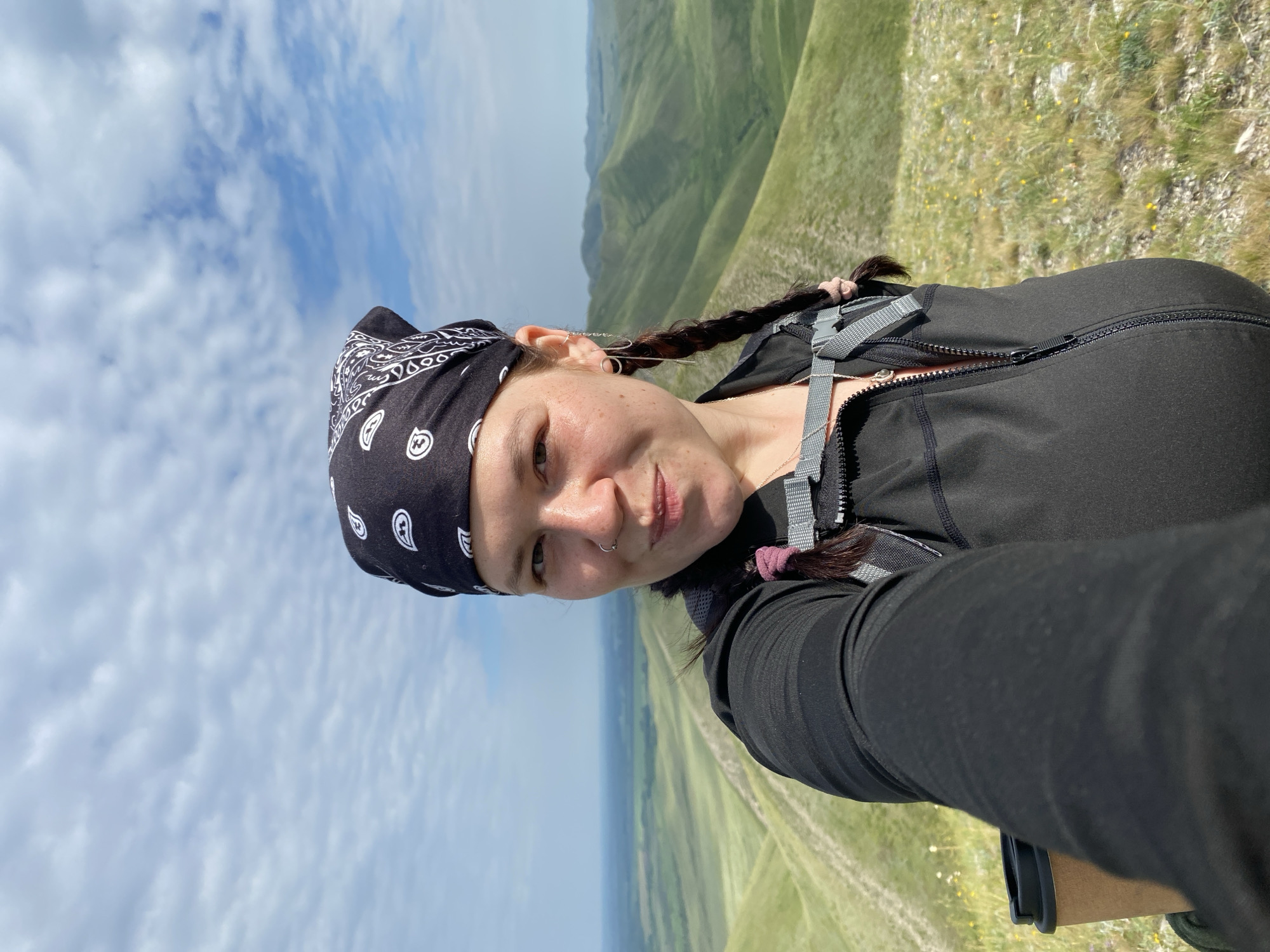 A woman with dark hair in two braids and a black bandana covering her head is taking a selfie on a grassy hilltop. She is wearing a black long-sleeved jacket, a backpack with grey straps, and a silver necklace. She has a nose ring and multiple earrings. The background shows rolling green hills under a partly cloudy sky, suggesting a rural or natural outdoor setting. This is a selfie taken in the vicinity of Verkhnyaya Chernorechka, Russia.
