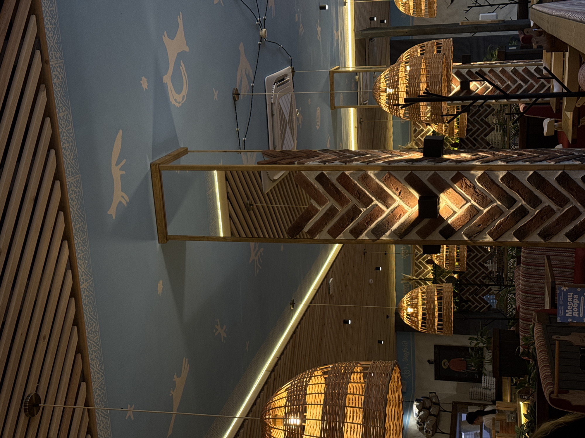The image captures an interior scene, likely a restaurant or cafe, in Mikhaylovsk, Russia. The ceiling is a prominent feature, painted light blue with white decorative patterns that appear to depict stylized animals, stars, and celestial bodies. Wooden slats run parallel across a portion of the ceiling. Hanging from the ceiling are multiple large, woven wicker pendant lamps with visible light bulbs, casting a warm glow.
In the foreground, a pillar clad in a herringbone pattern of reddish-brown bricks is visible on the right. Another similar pillar is seen further back on the left, also featuring the brickwork. These pillars create a sense of division and architectural interest within the space.
The environment includes seating areas with upholstered benches and chairs, some with red and white striped patterns. Tables are visible, suggesting dining or gathering spaces. In the background, elements of decor include what appears to be a painting or icon of a person in a hat, greenery in pots, and stacked items that could be tableware or decorative pieces. A woman with dark hair pulled back is partially visible on the left, seemingly attending to something behind a counter or service area. The overall atmosphere is warm and inviting, with a rustic yet decorative aesthetic.