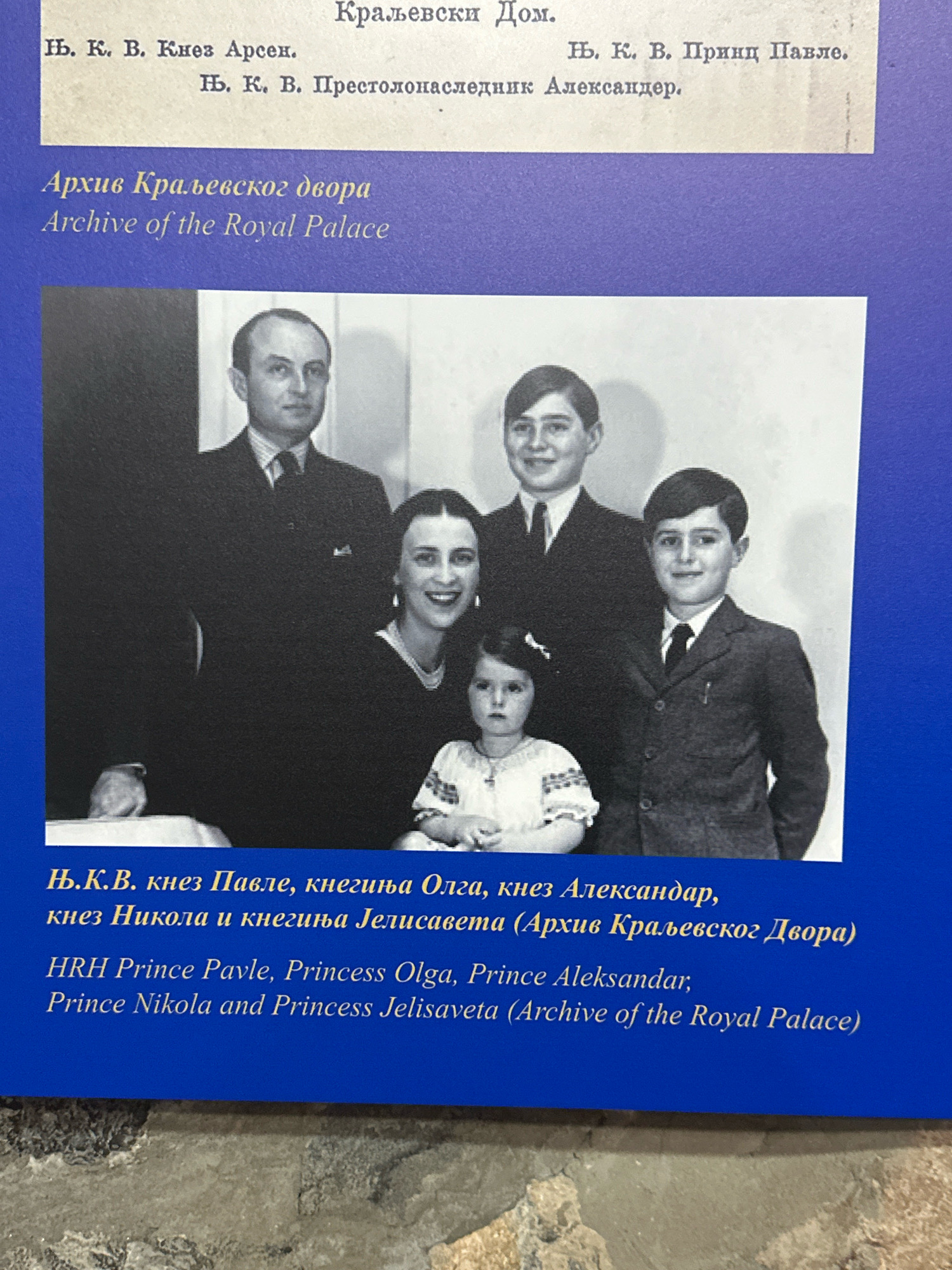 The image displays a formal portrait of a family group, likely taken in a studio setting, with a blue background. At the top, there are inscriptions in Cyrillic and Latin script, identifying the individuals and referencing the "Archive of the Royal Palace." The individuals depicted are Prince Pavle, Princess Olga, Prince Aleksandar, Princess Jelisaveta, and two other children. The portrait is presented as part of an exhibition or display, possibly at the Oplenac complex in Topola, Serbia, which houses the Royal Mausoleum and other royal residences. The inscription specifies that this is from the "Archive of the Royal Palace."