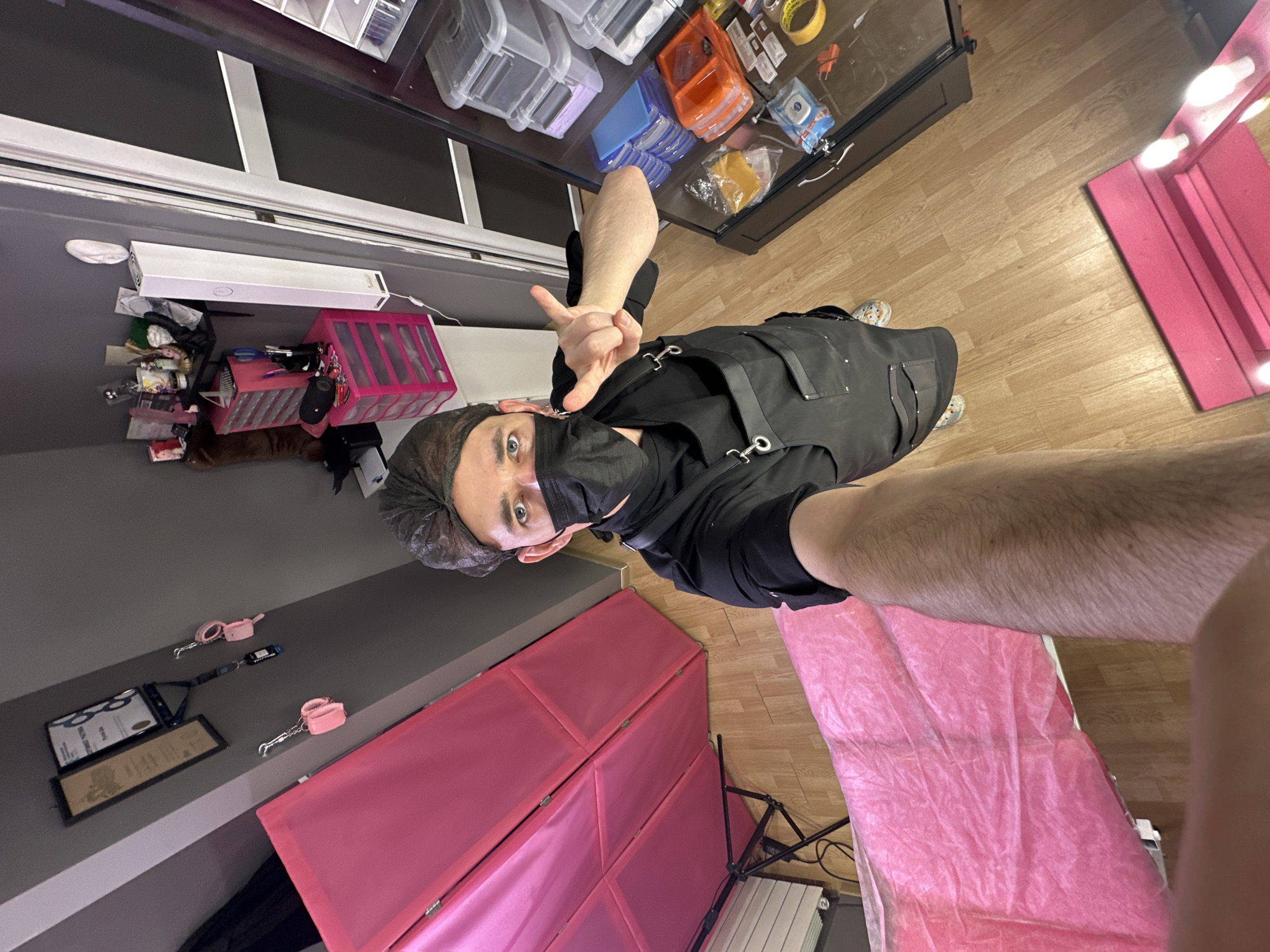 An overhead selfie depicts a man in a cosmetic or beauty studio located in Saratov, Russia. He wears a black short-sleeved shirt, a black apron with metallic fasteners, a black face mask, and a disposable hairnet; his blue eyes are visible above the mask. His right arm is extended, holding the camera, while his left hand gestures with the index and pinky fingers extended. The floor is light wood laminate. To the left, a pink folding screen stands next to a treatment table covered with a pink disposable sheet. On the gray wall, two framed documents are displayed, along with two pairs of pink faux fur handcuffs and a lanyard hanging from hooks. Beneath these are shelves holding various cosmetic products and two pink plastic drawer units containing tools and supplies. A white rectangular air purifier is affixed to the wall. To the right, a tall glass display cabinet holds multiple clear and colored plastic containers filled with professional-grade supplies. A pink vanity mirror with exposed bulbs is partially visible on the lower right floor.