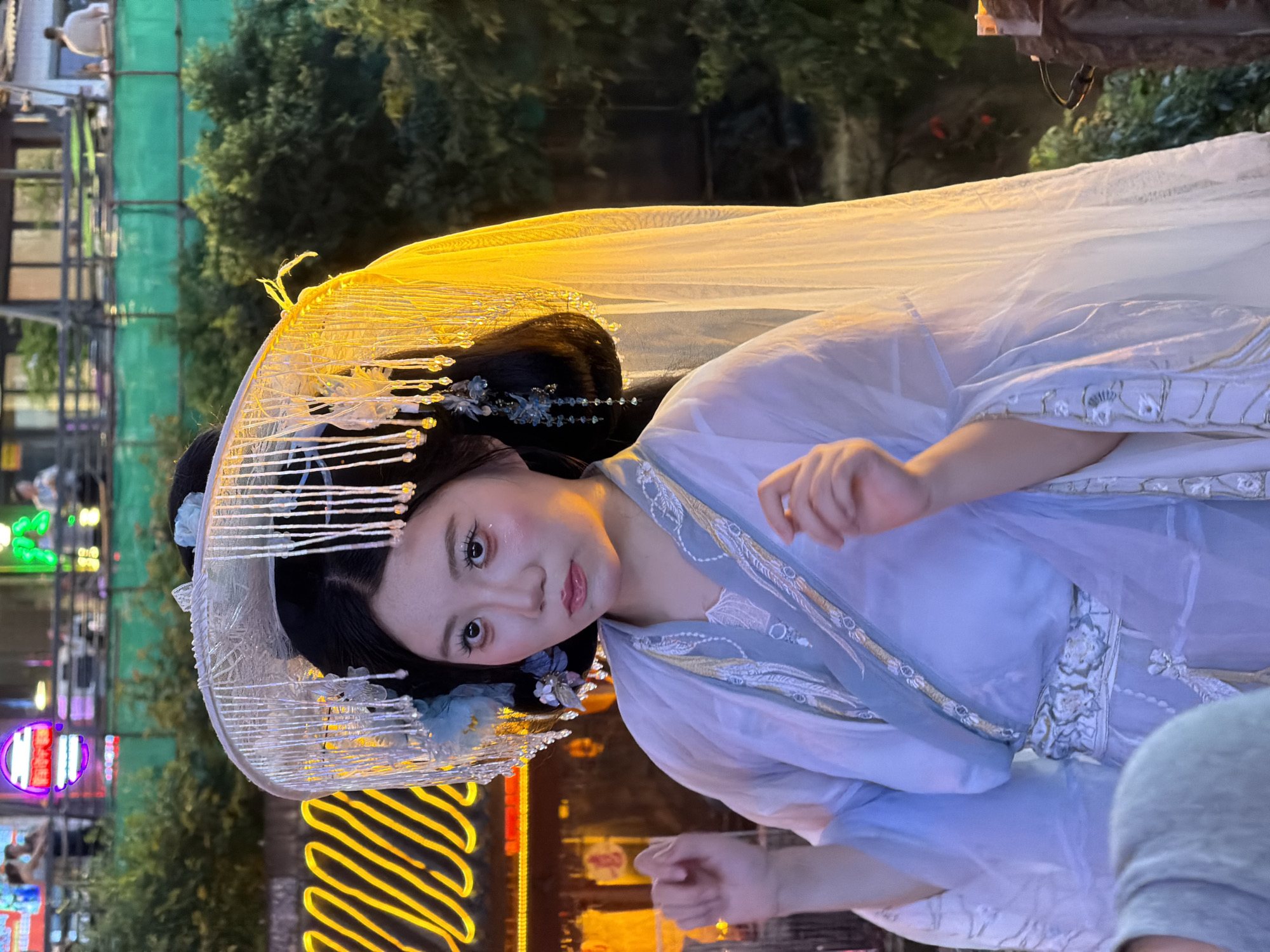 A person dressed in traditional Chinese attire, including a large, ornate hat adorned with beads and flowers, stands outdoors at night. The person wears a layered, light blue and white garment with intricate embroidery and a decorative belt. Their makeup is pronounced, with heavy eyeliner and blush. The background features blurred neon lights and greenery, suggesting an urban environment, possibly a tourist area or festival. The lighting is warm, highlighting the subject and casting a yellow glow on parts of the costume.