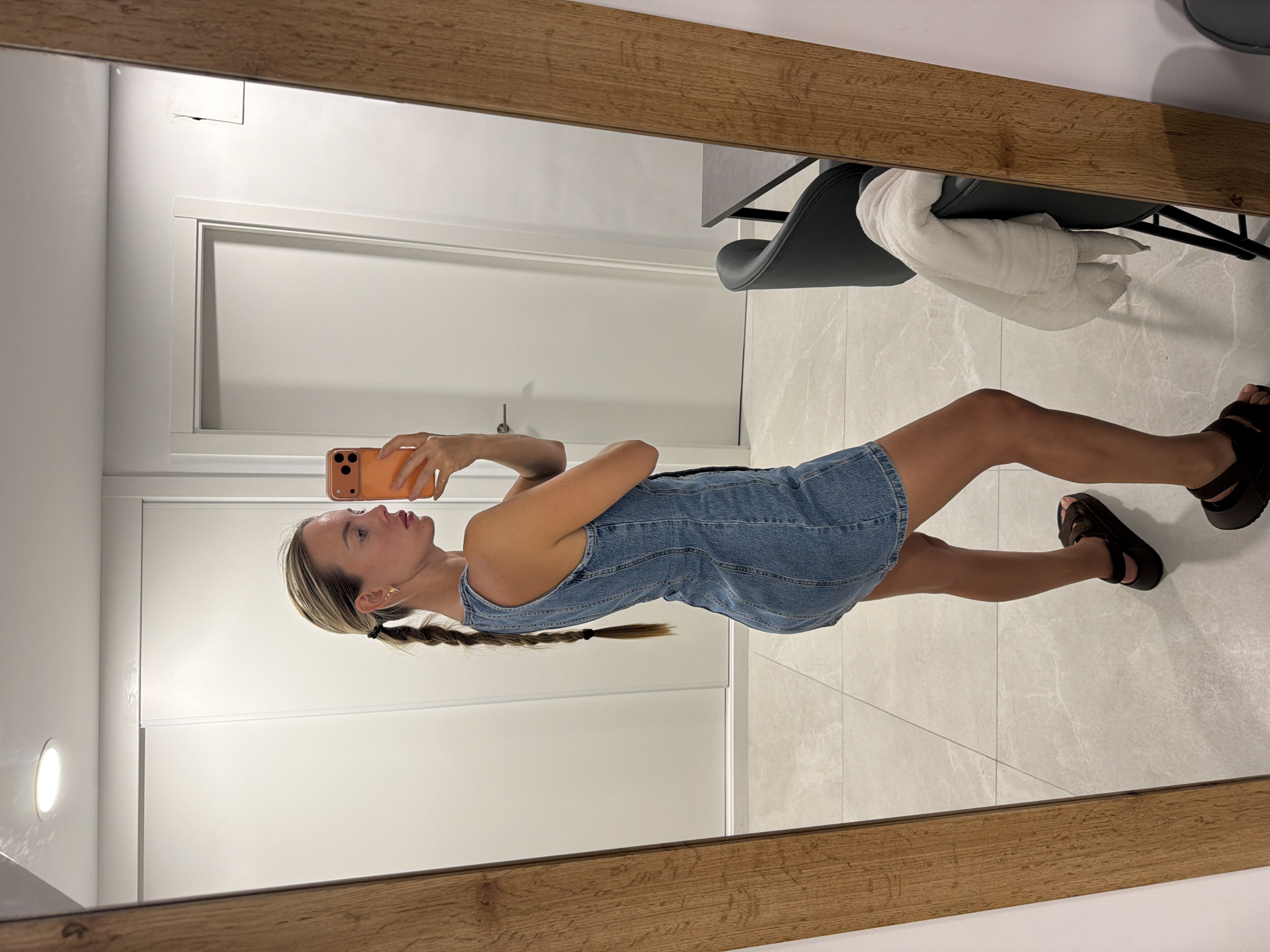 A young woman, seen from the side, stands in a brightly lit room and takes a selfie in a full-length mirror. She has blonde hair pulled back into a braid and is wearing a denim dress and dark sandals. The room appears to be a bedroom or dressing area, with sliding doors in the background and tiled flooring. A dark chair with a white towel draped over it is visible in the reflection to the right of the woman. The mirror has a thick, wooden frame. The woman is holding an orange smartphone in her right hand.