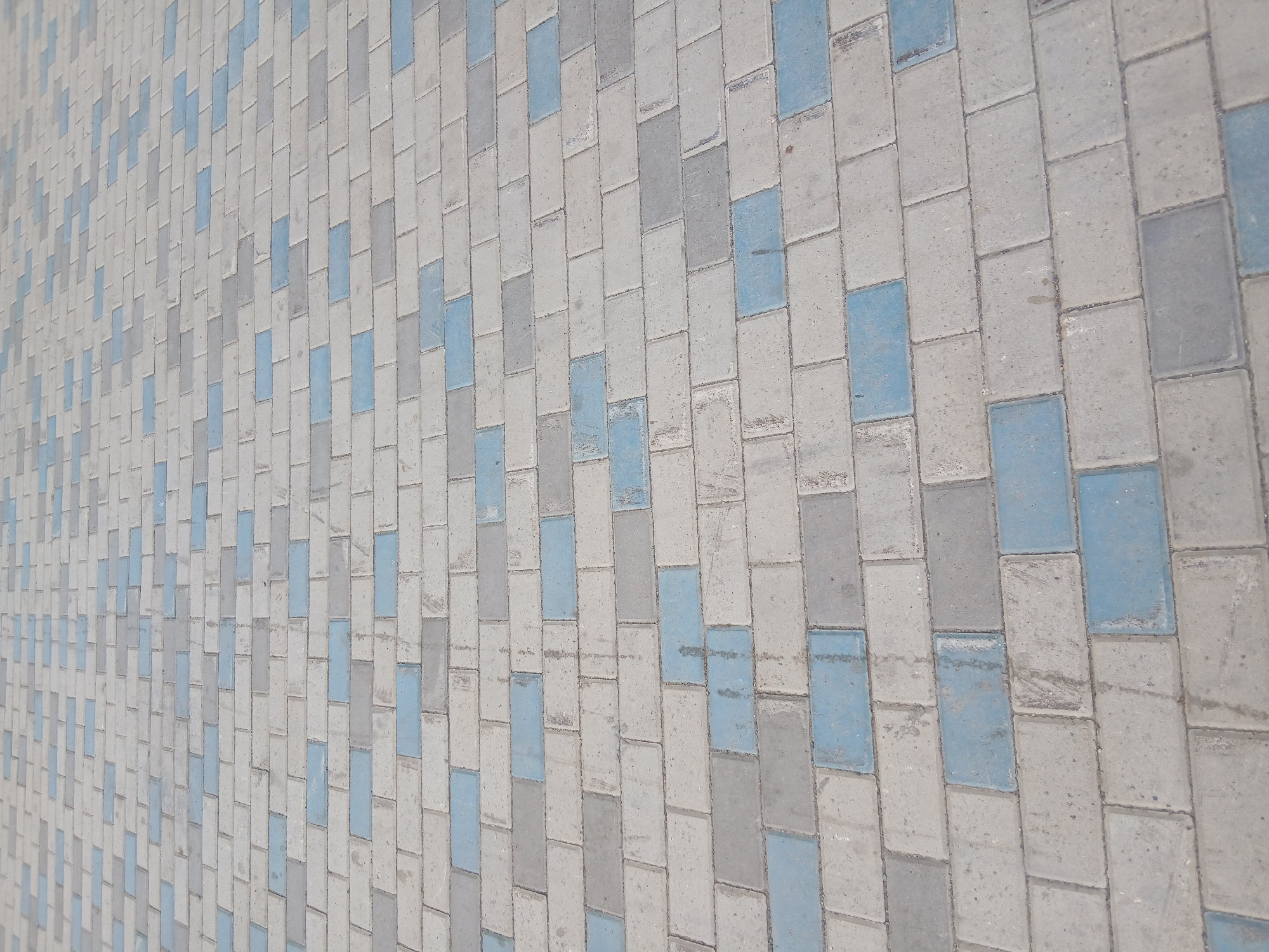 The image shows a section of a paved walkway in Astana, Kazakhstan. The walkway is constructed with rectangular interlocking pavers, a common material for public pedestrian areas. The pavers are of two distinct colors: a light gray and a muted blue. These colors are arranged in a repeating pattern, creating a visually interesting texture. The perspective of the image is from a slightly elevated viewpoint, looking down and across the walkway as it recedes into the distance. The pavers appear to be slightly worn and show some minor discoloration, suggesting they have been exposed to the elements and foot traffic. The scene is devoid of any people or other discernible objects, focusing solely on the geometric pattern and texture of the pavement.
