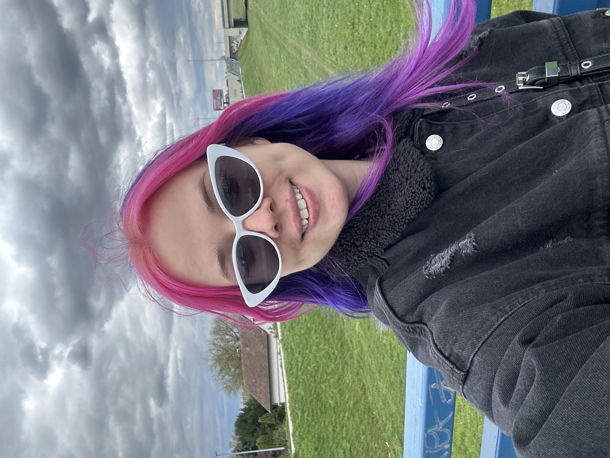 A medium shot shows a person with pink and purple hair, wearing white cat-eye sunglasses and a black denim jacket, smiling. The background includes a green field, a cloudy sky, a building with a brown roof, and a sign in the distance. The person is sitting on a blue structure with graffiti. The location is City of Zagreb, Croatia.