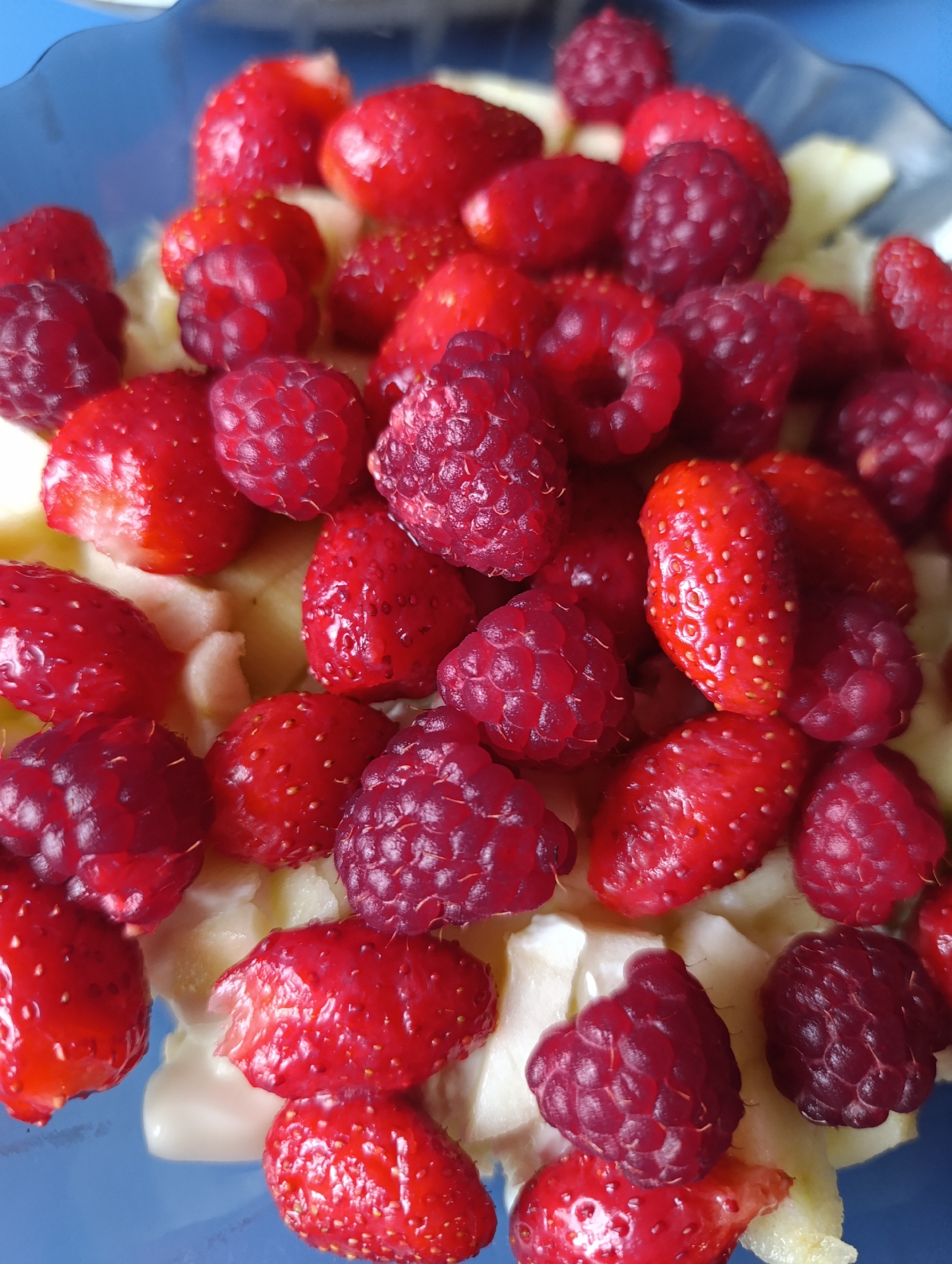 A close-up, overhead shot shows a blue bowl filled with a colorful fruit salad. The salad primarily consists of bright red strawberries and deep red raspberries, mixed with pieces of pale yellow fruit, likely pineapple or apple. The berries are whole and appear fresh, with visible seeds on the strawberries and the characteristic bumpy texture of raspberries. The lighting highlights the glossy surfaces of the fruit, creating a vibrant and appealing composition. The background shows a glimpse of the blue bowl and a soft, out-of-focus blue surface, possibly a table or tablecloth.