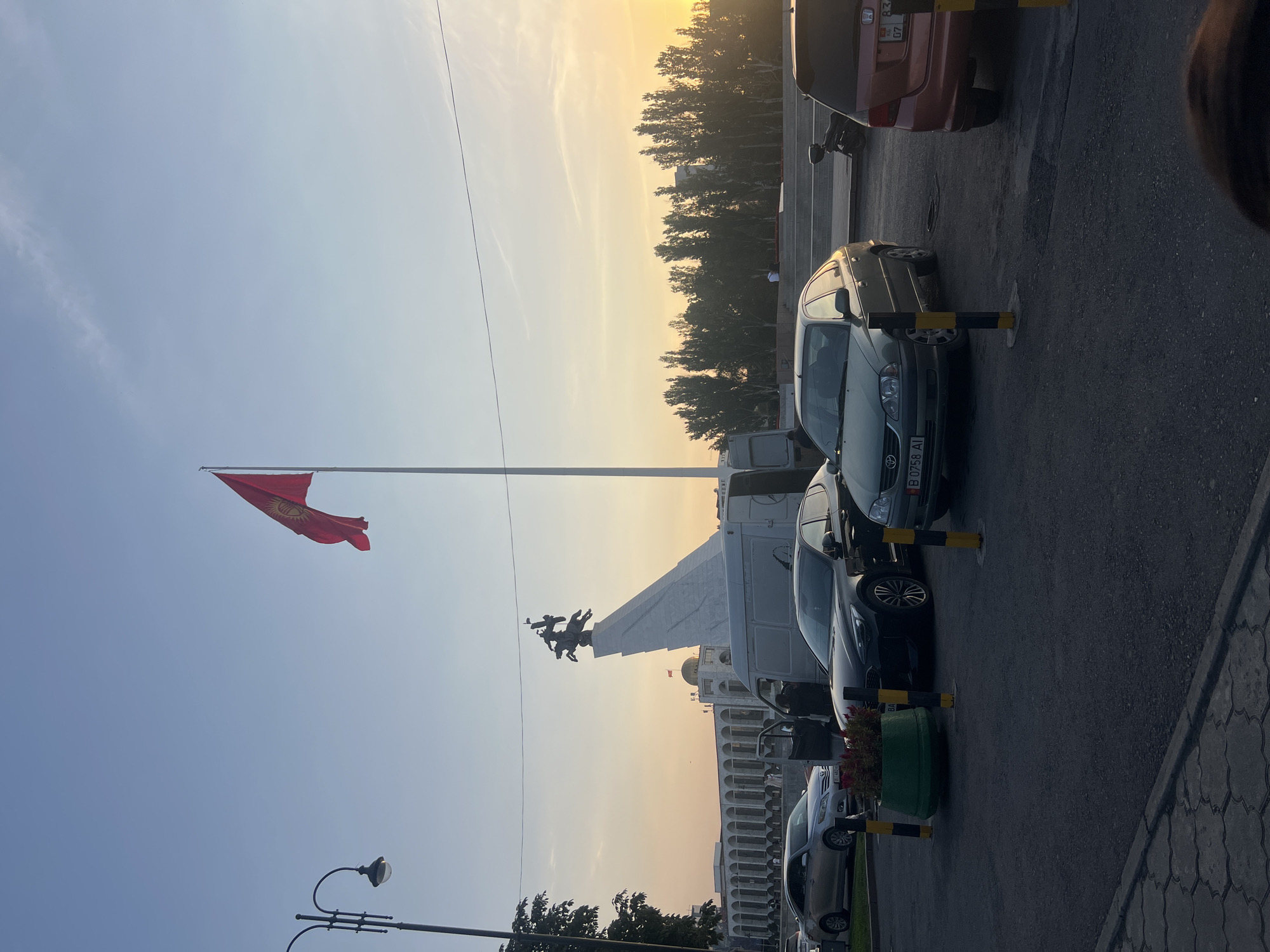The image depicts Ala-Too Square in Bishkek City, Kyrgyzstan, at sunset. In the foreground, several cars are parked, including a black Kia, a silver sedan, a green sedan, and a reddish-orange hatchback. A white van is parked with its driver's side door open, and a person is standing near it. A decorative planter with red flowers is situated between two black and yellow bollards. A lamppost is visible on the left side of the frame. In the background, the Manas Statue stands prominently on a white pedestal. Behind the statue, a building with a colonnade and a golden dome is visible, likely the State Historical Museum. A large red flag with a yellow sun symbol, representing the flag of Kyrgyzstan, flies from a tall flagpole. The sky is clear with a soft sunset glow. A line of tall trees borders the background, with buildings visible behind them. A motorcycle is parked near the reddish-orange hatchback. The overall scene conveys a calm, urban atmosphere during the evening hours.