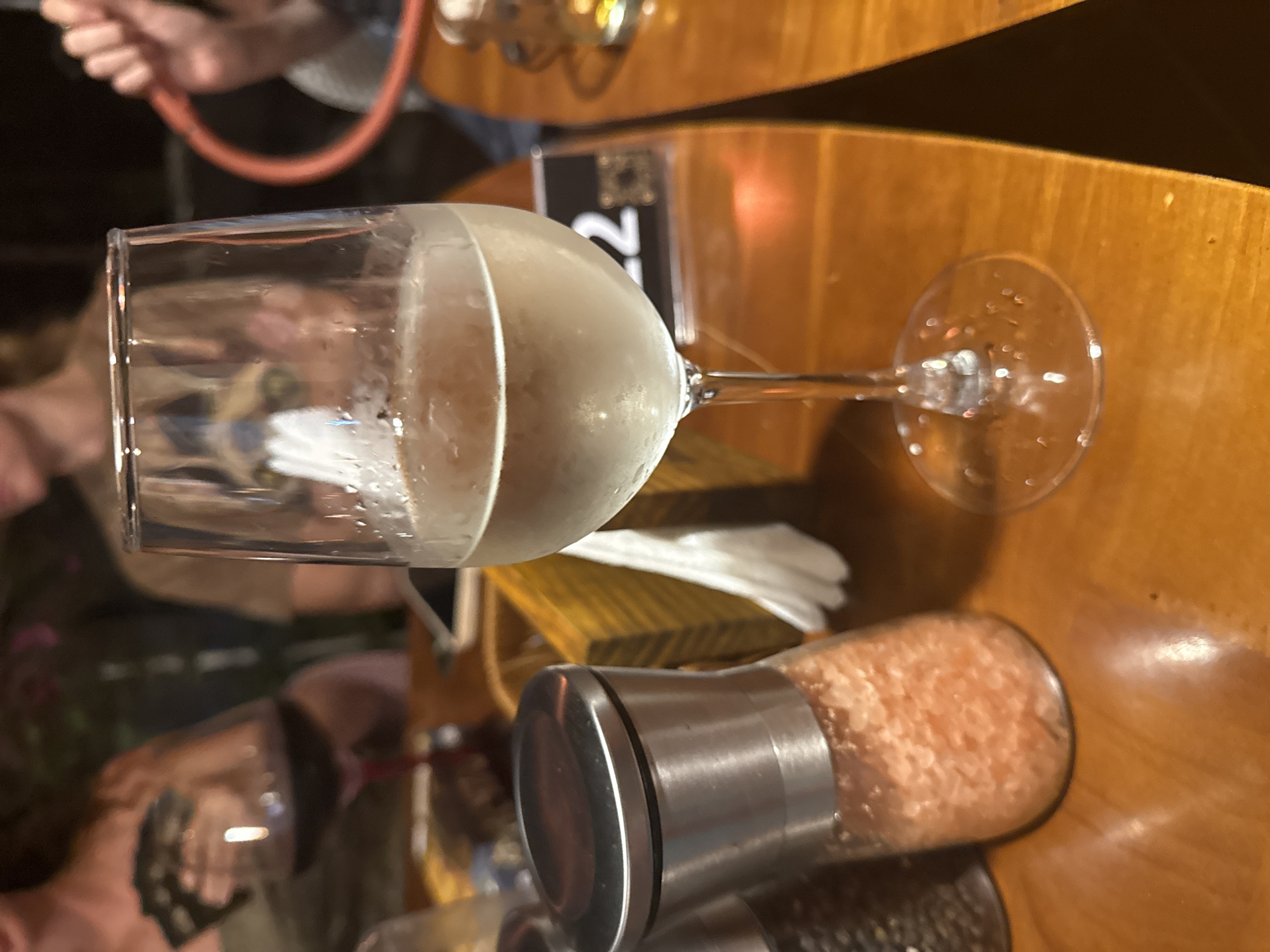 In the city of Bac Nha Trang Ward, Vietnam, a close-up shot focuses on a table at what appears to be an outdoor seating area. A glass of white wine, condensation forming on its surface, is the central subject, placed on a polished wooden table. To the left, a salt or pepper grinder with a metallic top is visible, filled with pink-hued granules. In the background, out of focus, two individuals are present. One is holding a glass of red wine, while the other is partially visible, with a reddish circular object, possibly part of a hookah, held in their hand. The lighting suggests it is evening or nighttime. A table number card with the digits "22" is also visible.
